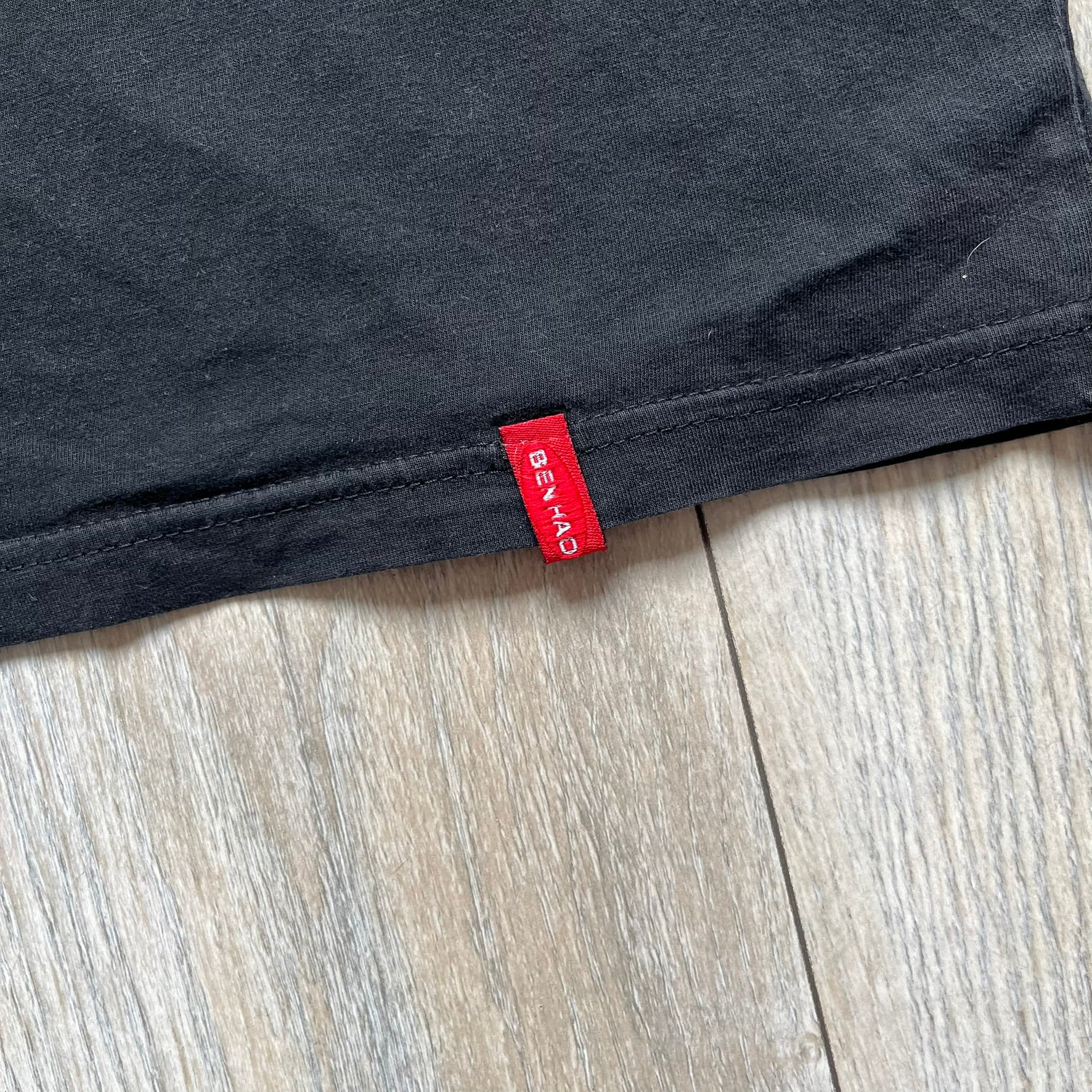 Counter-Strike 2000s Logo Tee is a black T-shirt featuring the classic Counter-Strike logo printed across the front. The design includes white “Counter” and “Strike” text separated by the armed character silhouette, with a red shadow layer behind the logo. A small red Ben Hao tag is attached near the bottom hem.