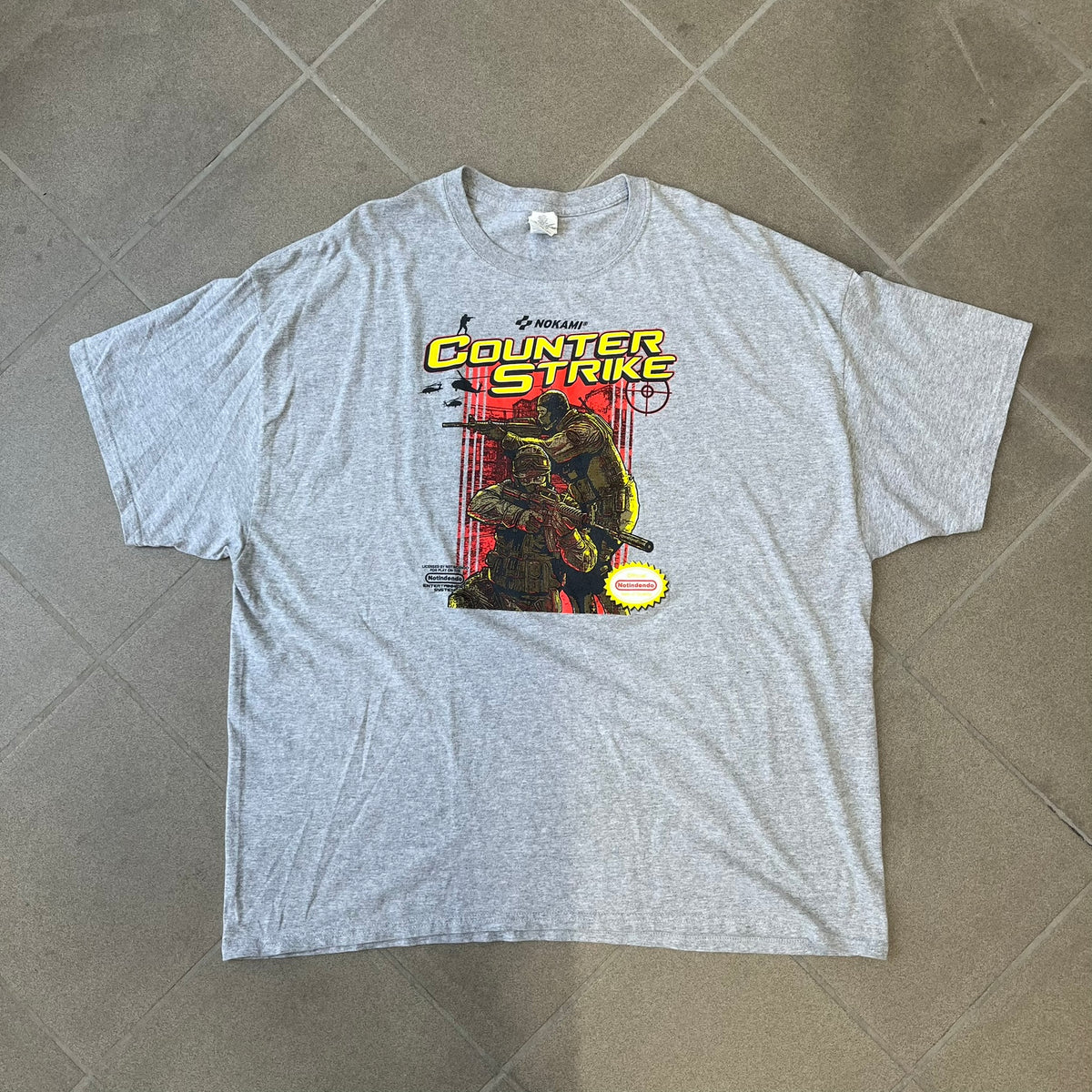 Counter Strike 2015 Arcade Block Gray Tee is a gray T-shirt featuring two Counter-Terrorists in yellow tones on a red background, with a yellow Counter-Strike text logo and a Nokami logo above the graphic. Two “Notindendo” logos sit on either side of the artwork. The shirt was available in the 2015 Arcade Block, specifically in the Nerd Block.