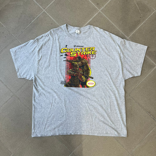 Counter Strike 2015 Arcade Block Gray Tee is a gray T-shirt featuring two Counter-Terrorists in yellow tones on a red background, with a yellow Counter-Strike text logo and a Nokami logo above the graphic. Two “Notindendo” logos sit on either side of the artwork. The shirt was available in the 2015 Arcade Block, specifically in the Nerd Block.