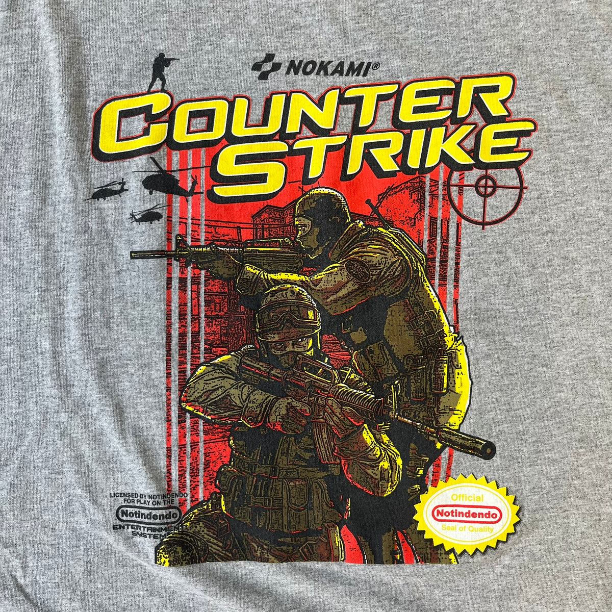 Counter Strike 2015 Arcade Block Gray Tee is a gray T-shirt featuring two Counter-Terrorists in yellow tones on a red background, with a yellow Counter-Strike text logo and a Nokami logo above the graphic. Two “Notindendo” logos sit on either side of the artwork. The shirt was available in the 2015 Arcade Block, specifically in the Nerd Block.