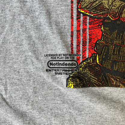 Counter Strike 2015 Arcade Block Gray Tee is a gray T-shirt featuring two Counter-Terrorists in yellow tones on a red background, with a yellow Counter-Strike text logo and a Nokami logo above the graphic. Two “Notindendo” logos sit on either side of the artwork. The shirt was available in the 2015 Arcade Block, specifically in the Nerd Block.