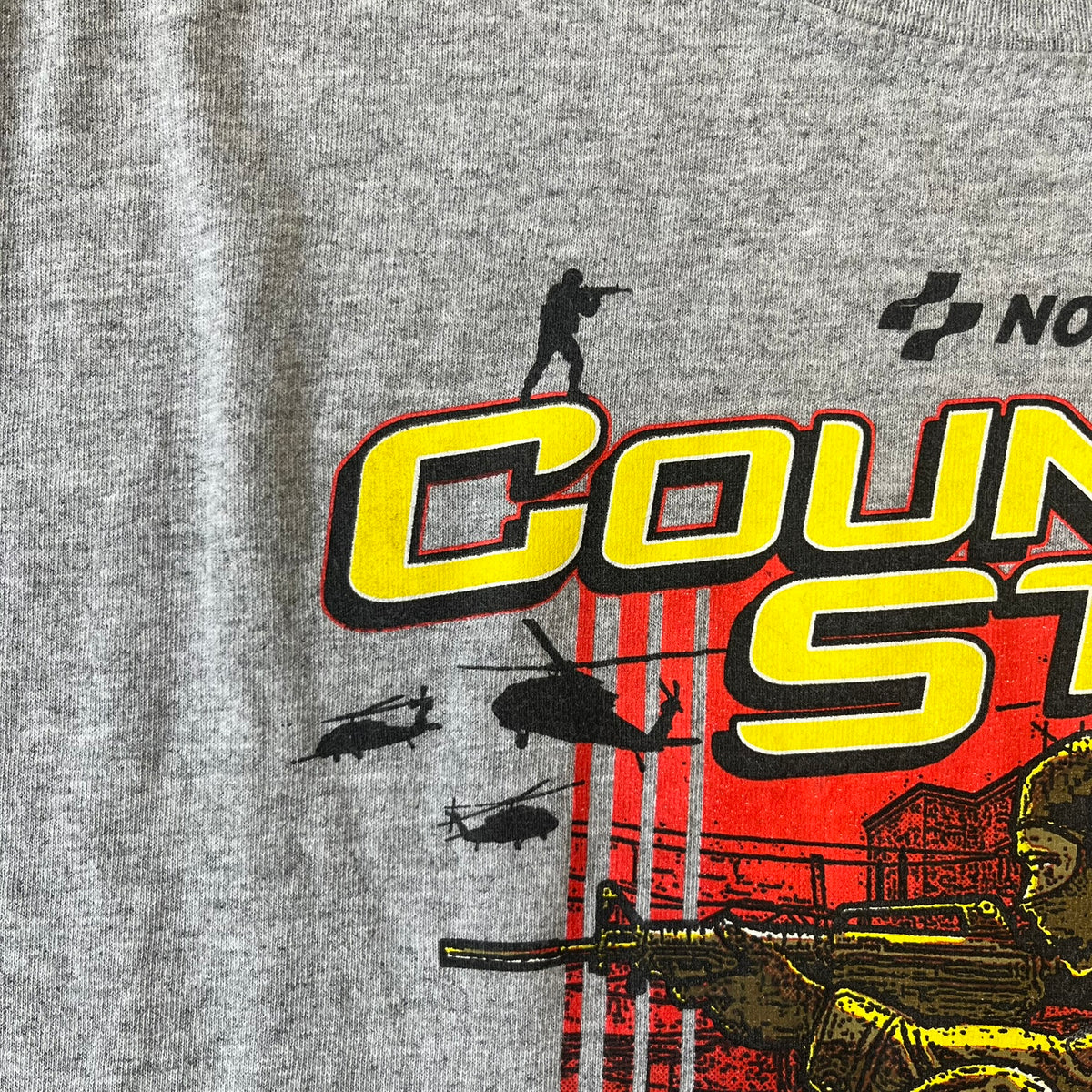 Counter Strike 2015 Arcade Block Gray Tee is a gray T-shirt featuring two Counter-Terrorists in yellow tones on a red background, with a yellow Counter-Strike text logo and a Nokami logo above the graphic. Two “Notindendo” logos sit on either side of the artwork. The shirt was available in the 2015 Arcade Block, specifically in the Nerd Block.