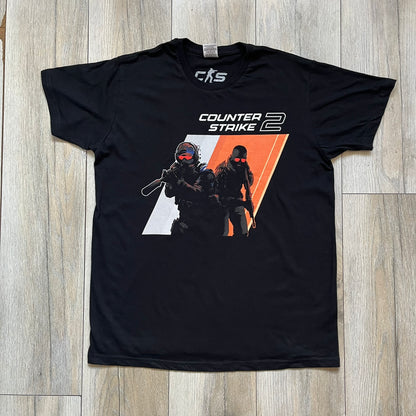 Counter-Strike 2 Promo 2023 Tee is a black T-shirt featuring the game cover graphic on the front, with the Counter-Strike 2 logo above two characters (terrorist and counter-terrorist) set against a white and orange background. The shirt includes a printed CS2 neck tag with 2023 Valve licensing.