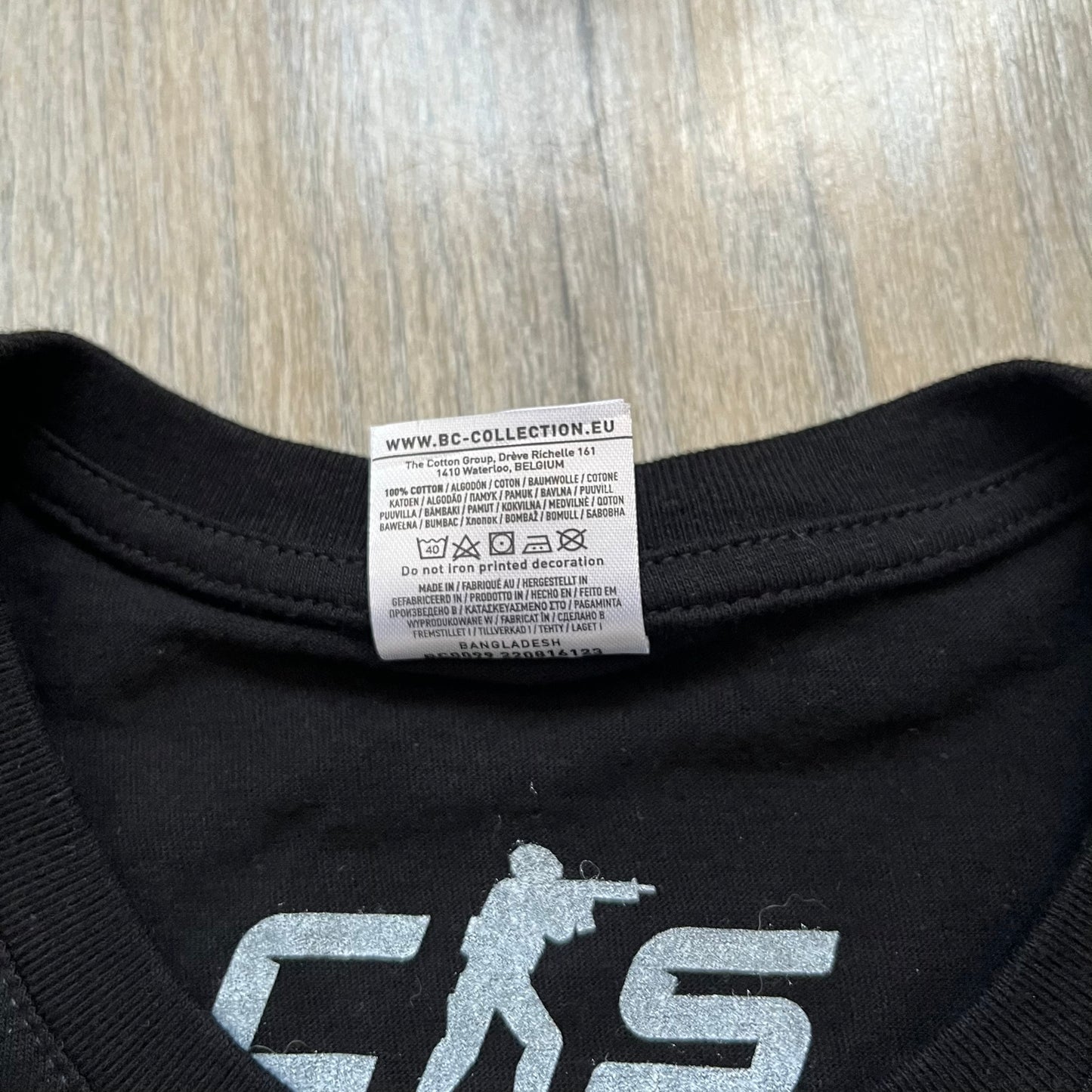 Counter-Strike 2 Promo 2023 Tee is a black T-shirt featuring the game cover graphic on the front, with the Counter-Strike 2 logo above two characters (terrorist and counter-terrorist) set against a white and orange background. The shirt includes a printed CS2 neck tag with 2023 Valve licensing.