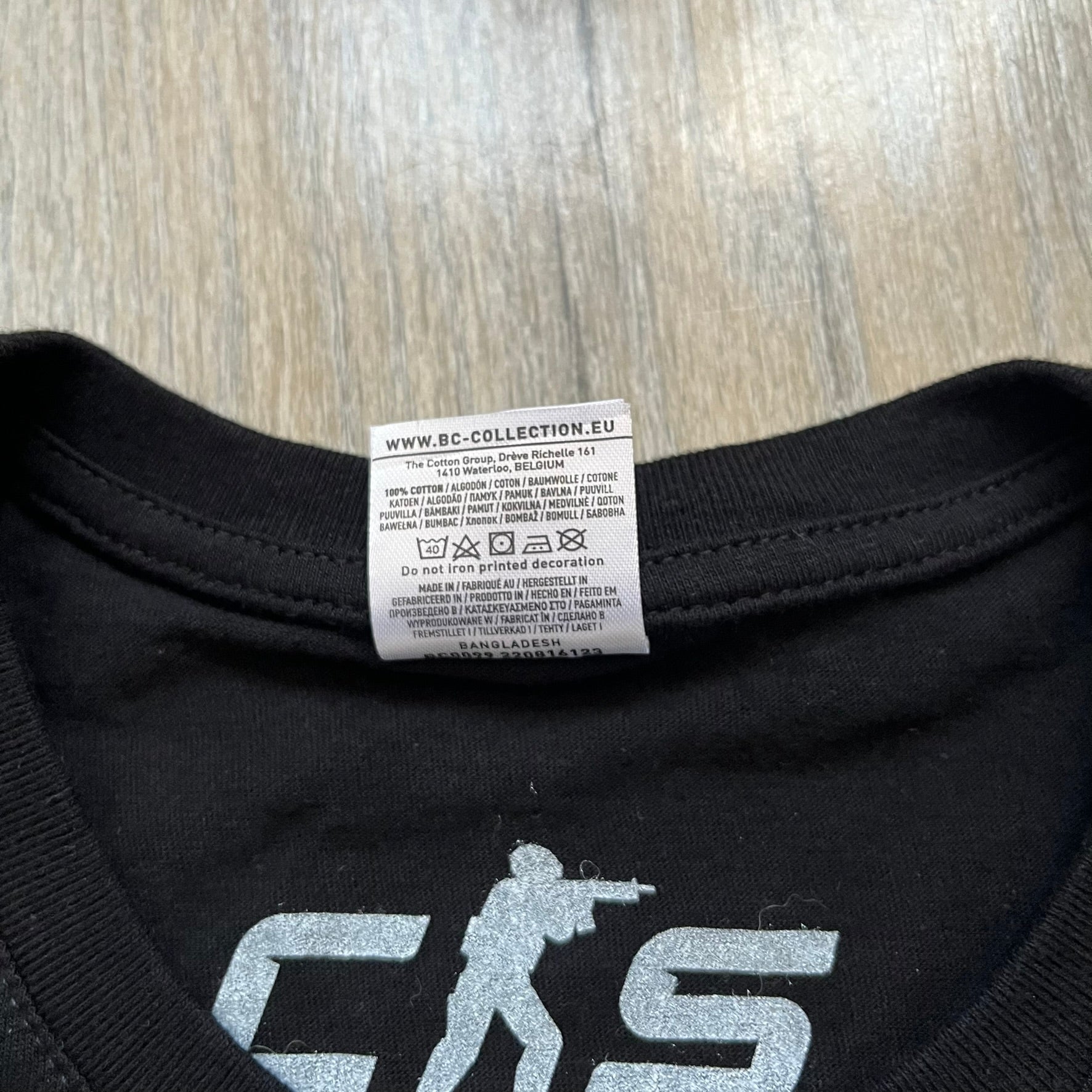 Counter-Strike 2 Promo 2023 Tee is a black T-shirt featuring the game cover graphic on the front, with the Counter-Strike 2 logo above two characters (terrorist and counter-terrorist) set against a white and orange background. The shirt includes a printed CS2 neck tag with 2023 Valve licensing.