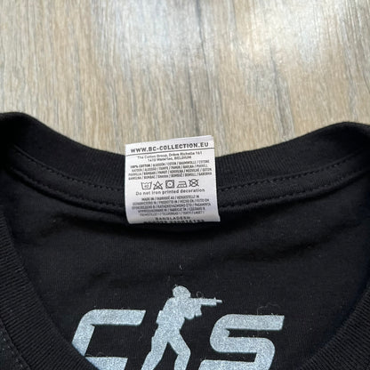 Counter-Strike 2 Promo 2023 Tee is a black T-shirt featuring the game cover graphic on the front, with the Counter-Strike 2 logo above two characters (terrorist and counter-terrorist) set against a white and orange background. The shirt includes a printed CS2 neck tag with 2023 Valve licensing.