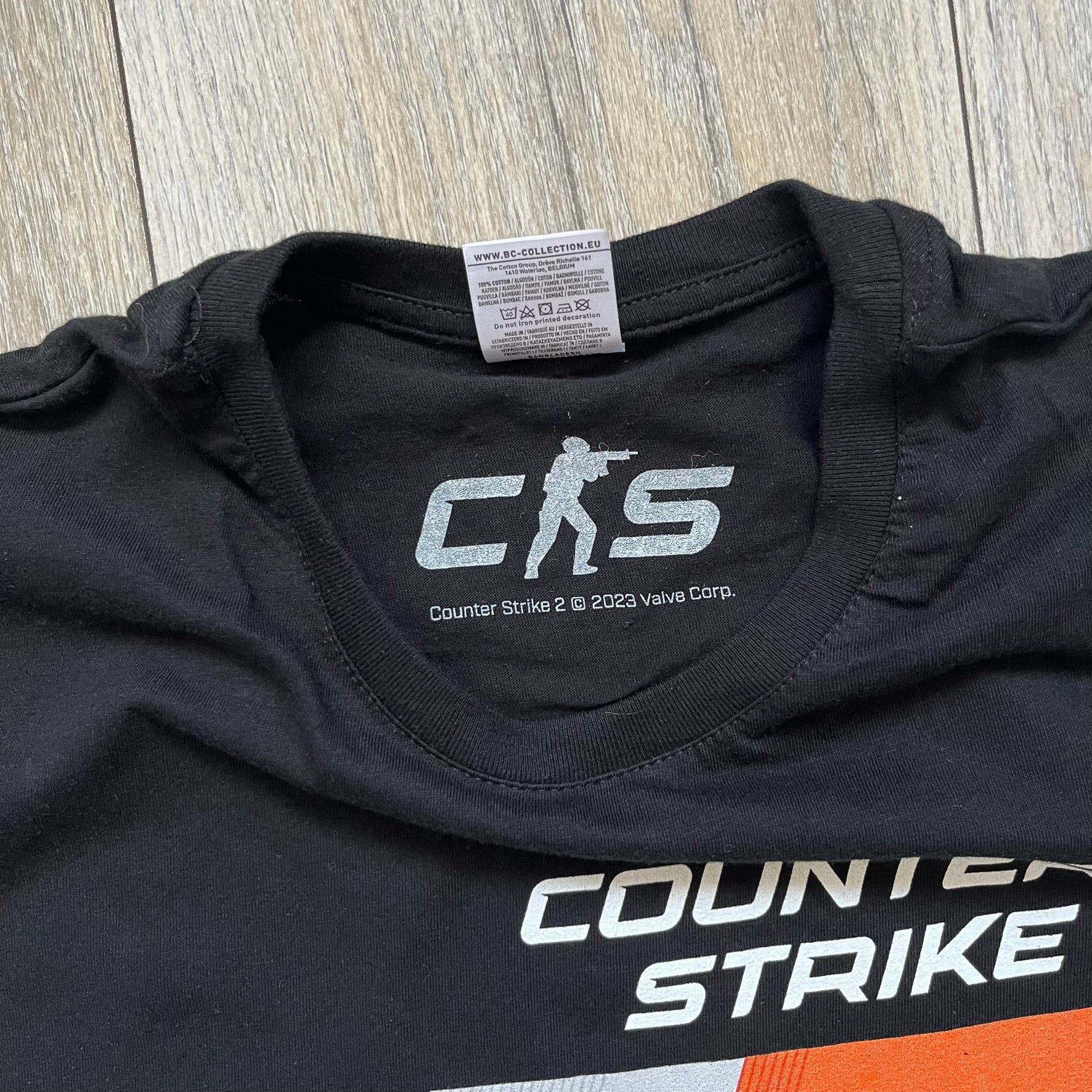 Counter-Strike 2 Promo 2023 Tee is a black T-shirt featuring the game cover graphic on the front, with the Counter-Strike 2 logo above two characters (terrorist and counter-terrorist) set against a white and orange background. The shirt includes a printed CS2 neck tag with 2023 Valve licensing.
