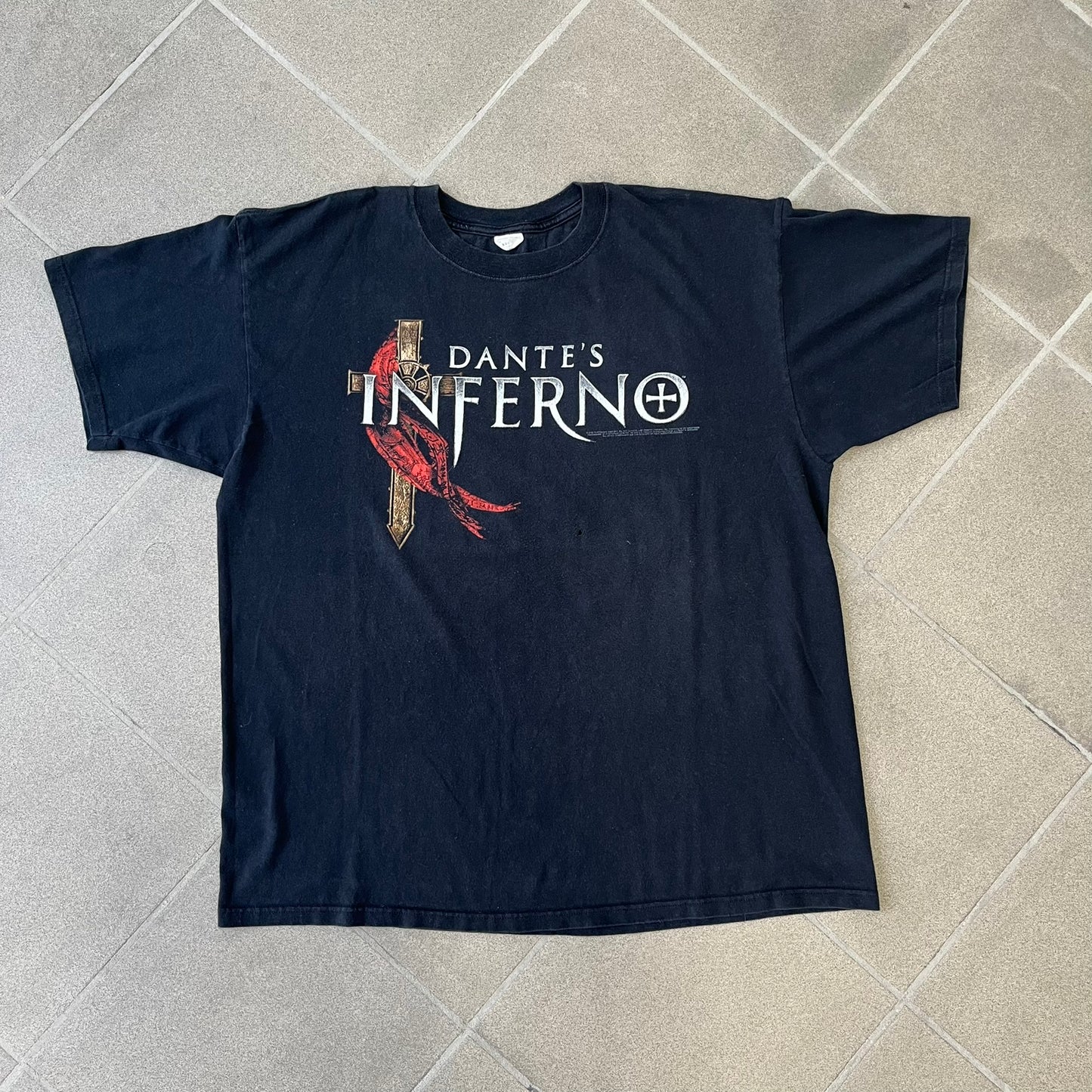 Dante's Inferno 2010 Black Promo Tee features a black construction made on an Anvil blank. The front has the Dante's Inferno game logo with a cross graphic on the left, featuring a red fabric-like detail, with small white licensing text underneath.

