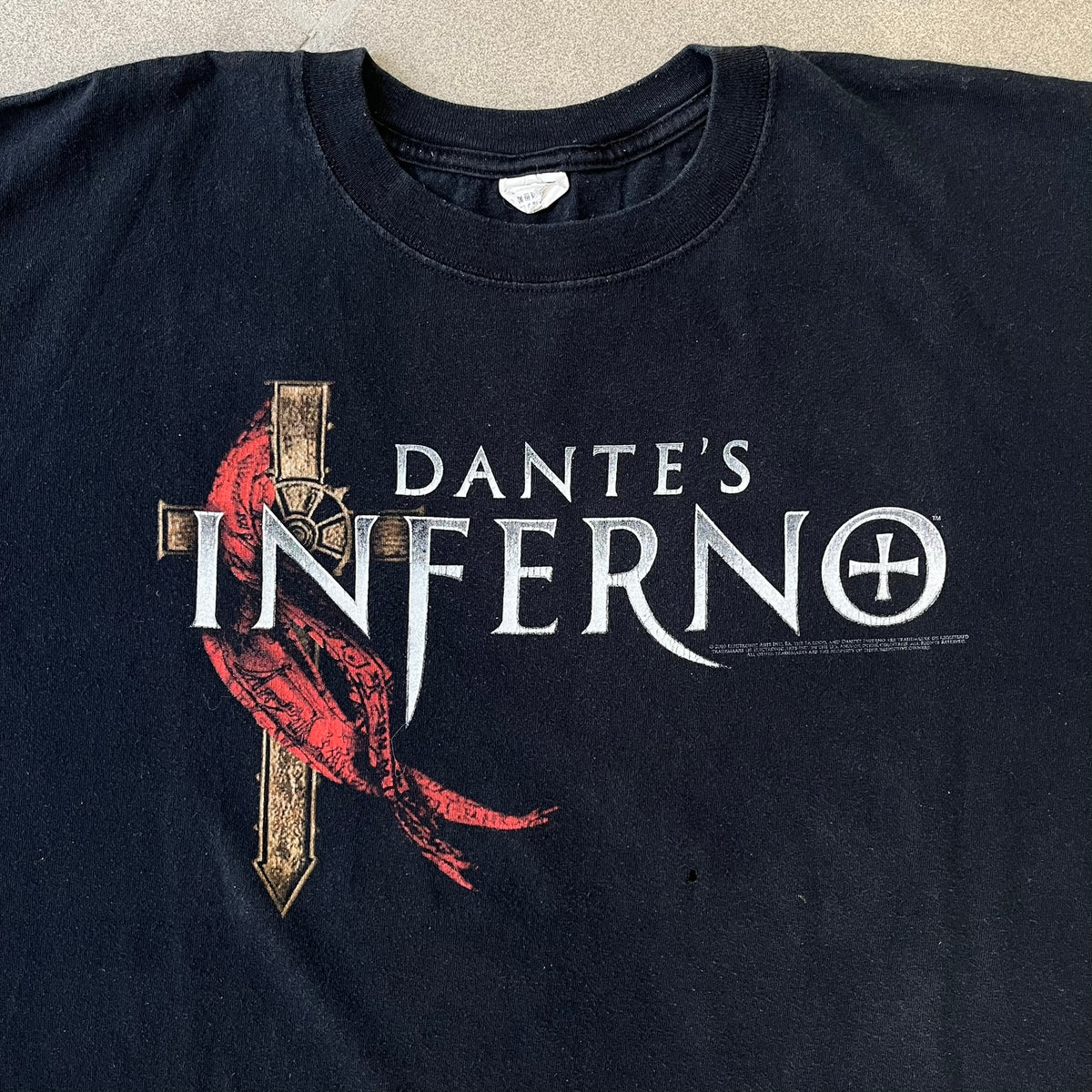 Dante's Inferno 2010 Black Promo Tee features a black construction made on an Anvil blank. The front has the Dante's Inferno game logo with a cross graphic on the left, featuring a red fabric-like detail, with small white licensing text underneath.
