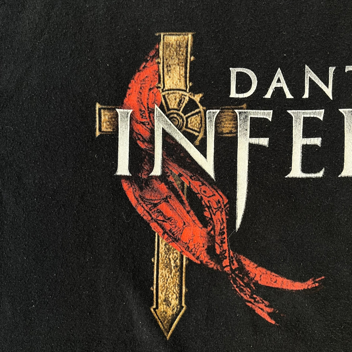 Dante's Inferno 2010 Black Promo Tee features a black construction made on an Anvil blank. The front has the Dante's Inferno game logo with a cross graphic on the left, featuring a red fabric-like detail, with small white licensing text underneath.
