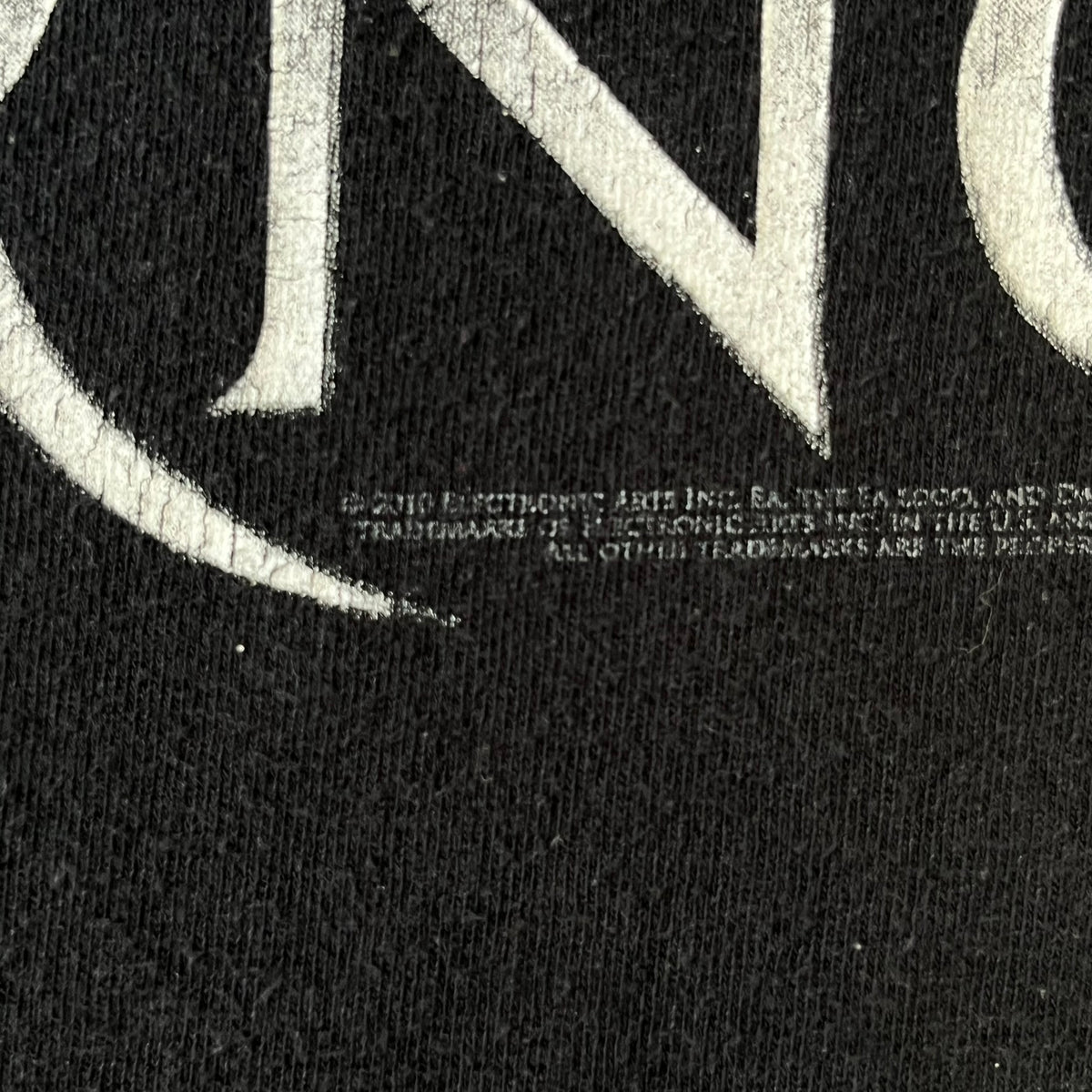 Dante's Inferno 2010 Black Promo Tee features a black construction made on an Anvil blank. The front has the Dante's Inferno game logo with a cross graphic on the left, featuring a red fabric-like detail, with small white licensing text underneath.
