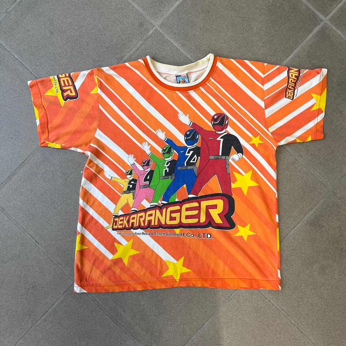 Dekaranger 2004 Orange All Over Print Tee is an orange all-over print T-shirt featuring the same design on both front and back, showing five Dekarangers lined up with "Dekaranger" text and licensing below, set against a background of white stripes and yellow stars. Dekaranger branding appears on both sleeves, and it carries a Gaoranger neck tag.