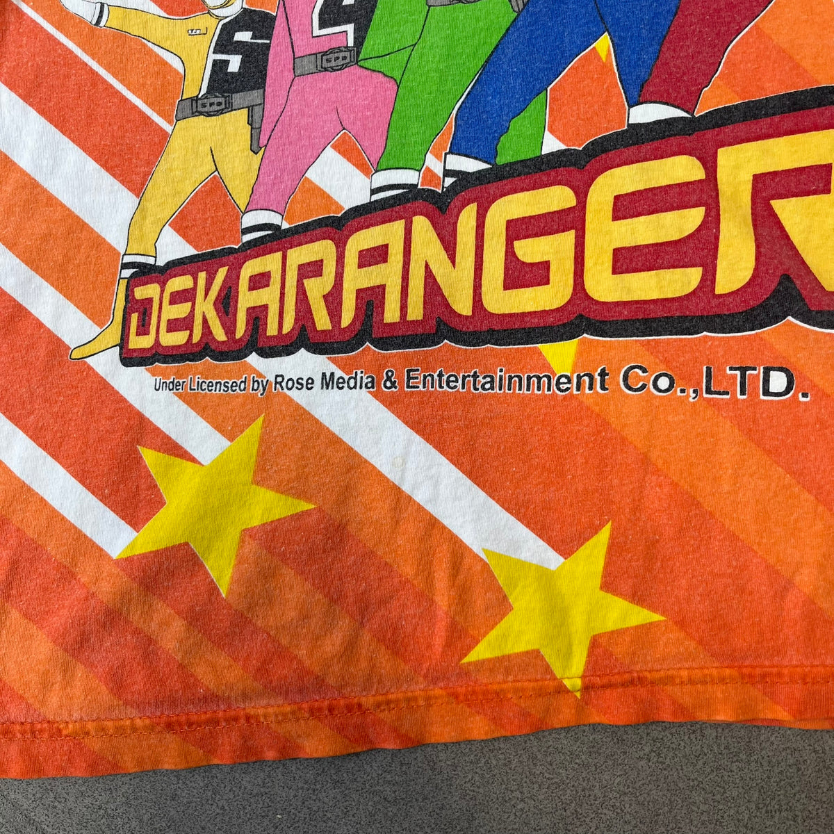 Dekaranger 2004 Orange All Over Print Tee is an orange all-over print T-shirt featuring the same design on both front and back, showing five Dekarangers lined up with "Dekaranger" text and licensing below, set against a background of white stripes and yellow stars. Dekaranger branding appears on both sleeves, and it carries a Gaoranger neck tag.