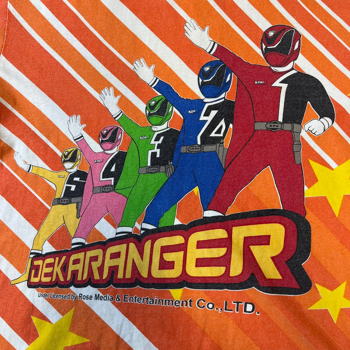 Dekaranger 2004 Orange All Over Print Tee is an orange all-over print T-shirt featuring the same design on both front and back, showing five Dekarangers lined up with "Dekaranger" text and licensing below, set against a background of white stripes and yellow stars. Dekaranger branding appears on both sleeves, and it carries a Gaoranger neck tag.