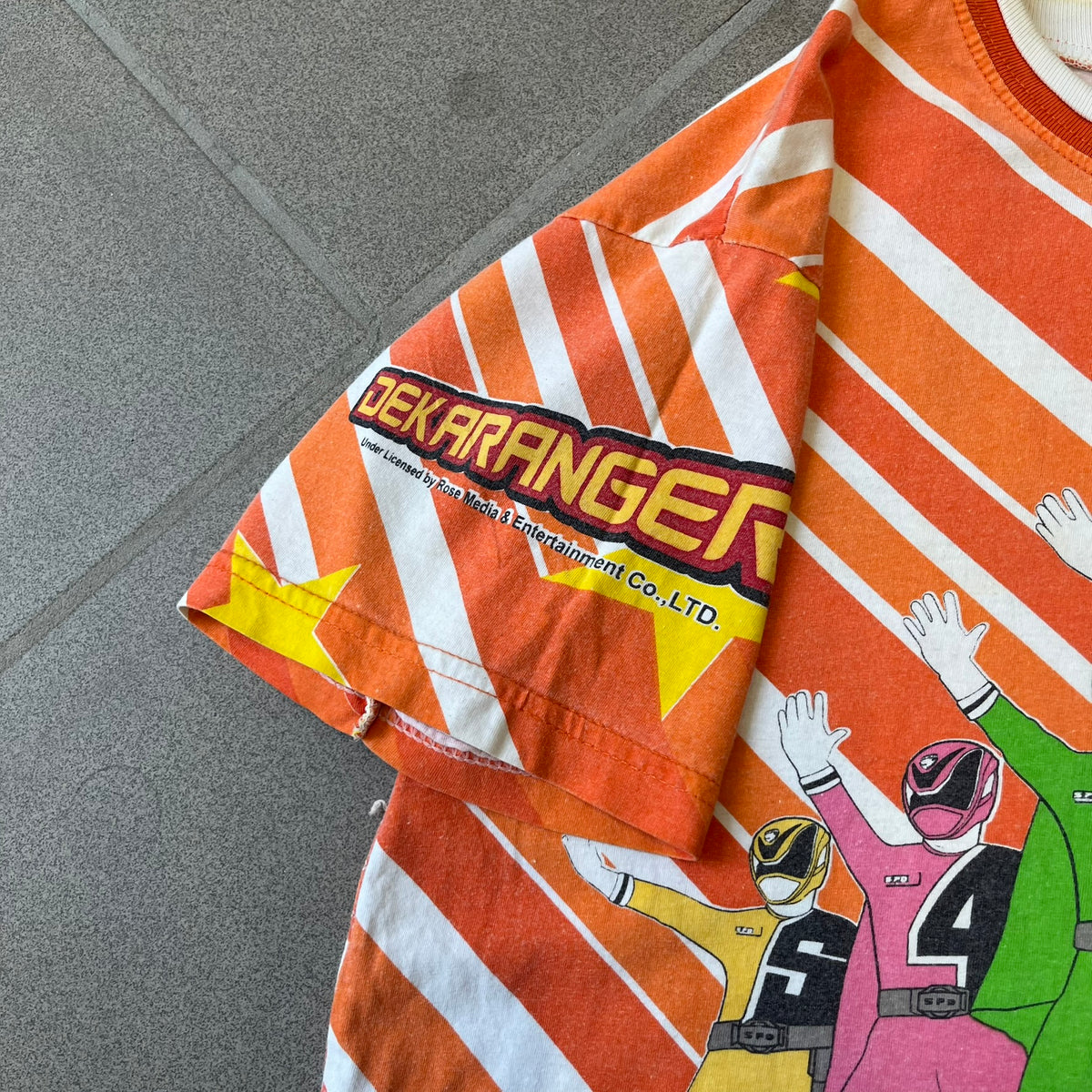 Dekaranger 2004 Orange All Over Print Tee is an orange all-over print T-shirt featuring the same design on both front and back, showing five Dekarangers lined up with "Dekaranger" text and licensing below, set against a background of white stripes and yellow stars. Dekaranger branding appears on both sleeves, and it carries a Gaoranger neck tag.