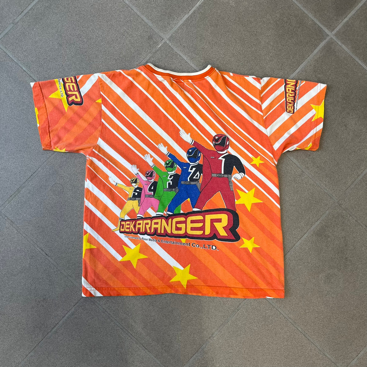 Dekaranger 2004 Orange All Over Print Tee is an orange all-over print T-shirt featuring the same design on both front and back, showing five Dekarangers lined up with "Dekaranger" text and licensing below, set against a background of white stripes and yellow stars. Dekaranger branding appears on both sleeves, and it carries a Gaoranger neck tag.