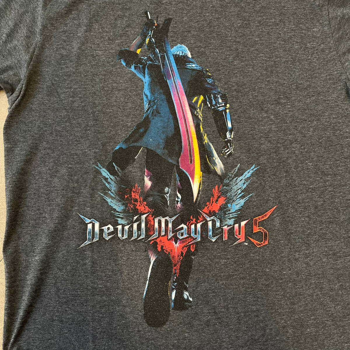 Devil May Cry 5 Promo 2019 Gray Tee is made on a Gildan blank in grey cotton. The front features the Devil May Cry 5 game cover but only with Nero holding his sword, along with large “Devil May Cry 5” text. Made as a promotional T-shirt for the game.