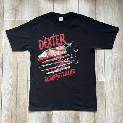 Dexter “Blood Never Lies” 2017 Tee is a black T-shirt featuring a front graphic with red blood-style “Dexter” and “Blood Never Lies” text. Between the texts, the print shows blood-covered tools with Dexter visible in the reflection of a saw. Small 2017 Showtime licensing text appears beneath the graphic, confirming official series merchandise.