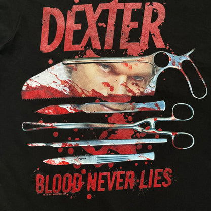 Dexter “Blood Never Lies” 2017 Tee is a black T-shirt featuring a front graphic with red blood-style “Dexter” and “Blood Never Lies” text. Between the texts, the print shows blood-covered tools with Dexter visible in the reflection of a saw. Small 2017 Showtime licensing text appears beneath the graphic, confirming official series merchandise.