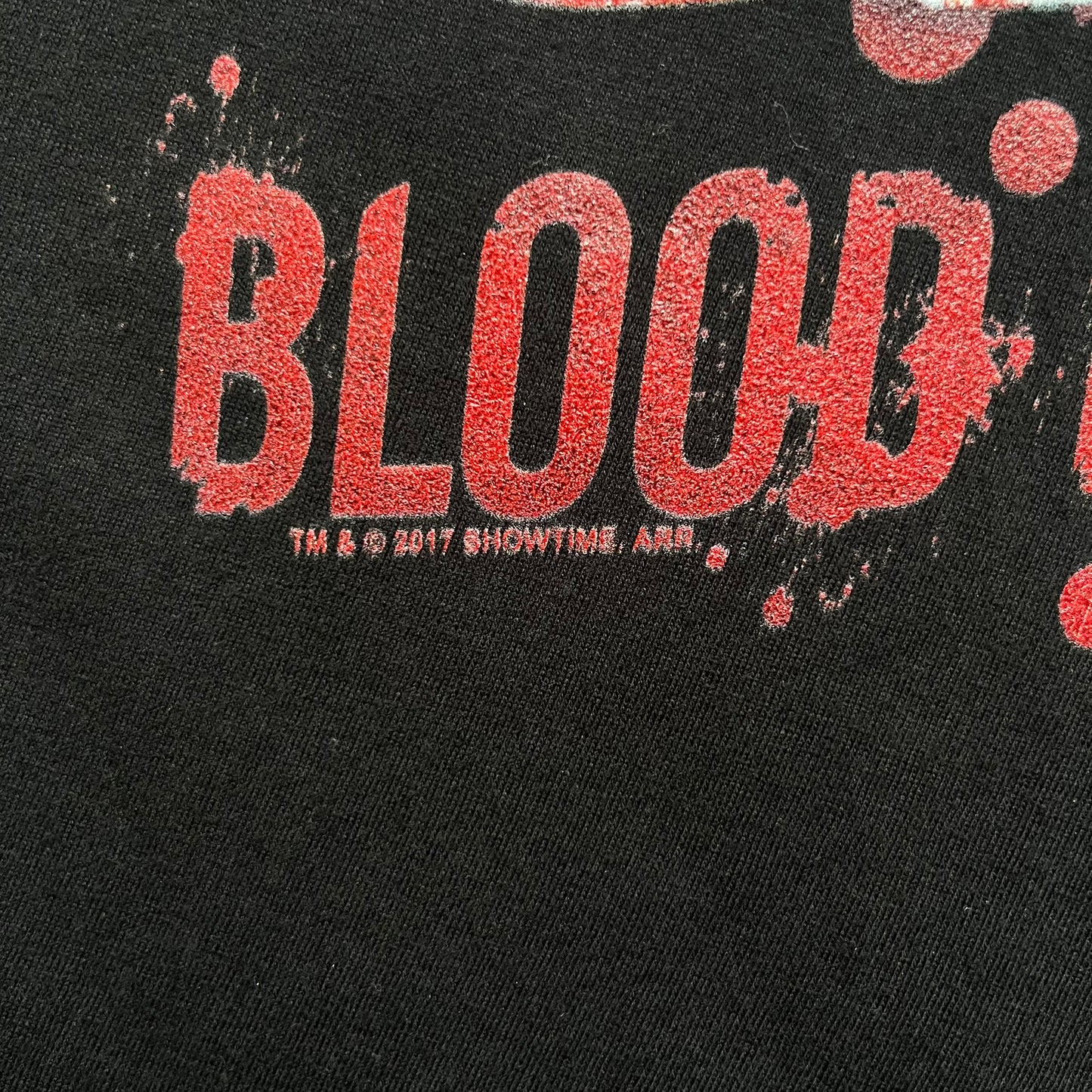 Dexter “Blood Never Lies” 2017 Tee is a black T-shirt featuring a front graphic with red blood-style “Dexter” and “Blood Never Lies” text. Between the texts, the print shows blood-covered tools with Dexter visible in the reflection of a saw. Small 2017 Showtime licensing text appears beneath the graphic, confirming official series merchandise.