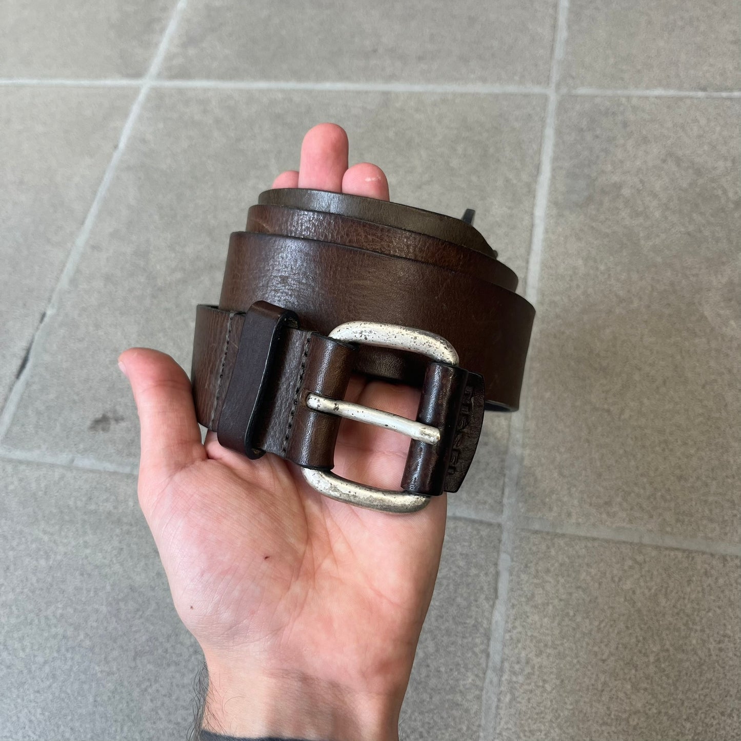 Diesel 2000's Brown Leather Belt features a brown 100% cow leather construction with a simple silver-tone buckle. A small leather piece with Diesel text is attached to the buckle and rotates freely. The belt is marked “Extra Tough,” with gold Diesel text printed on the inside. Made in Italy, with an additional attached section at the end for adjusting the length.
