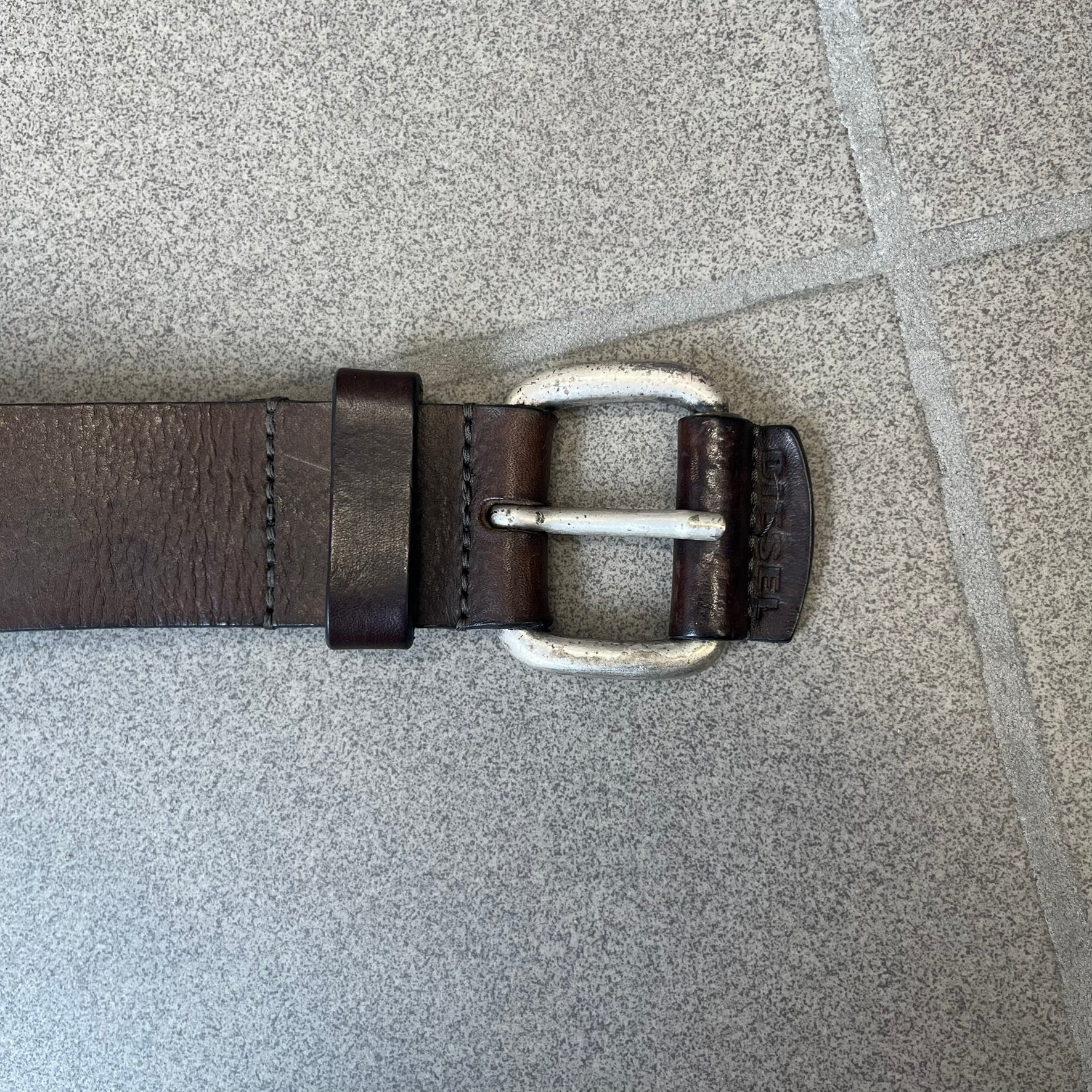 Diesel 2000's Brown Leather Belt features a brown 100% cow leather construction with a simple silver-tone buckle. A small leather piece with Diesel text is attached to the buckle and rotates freely. The belt is marked “Extra Tough,” with gold Diesel text printed on the inside. Made in Italy, with an additional attached section at the end for adjusting the length.
