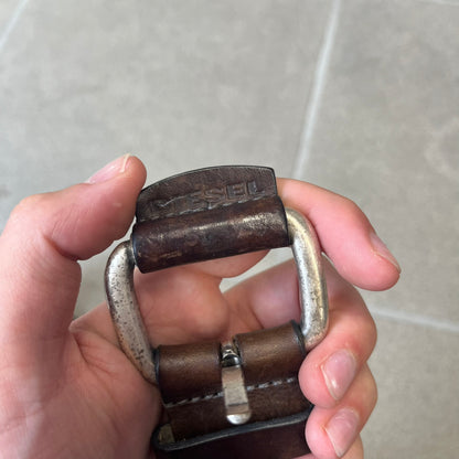 Diesel 2000's Brown Leather Belt features a brown 100% cow leather construction with a simple silver-tone buckle. A small leather piece with Diesel text is attached to the buckle and rotates freely. The belt is marked “Extra Tough,” with gold Diesel text printed on the inside. Made in Italy, with an additional attached section at the end for adjusting the length.
