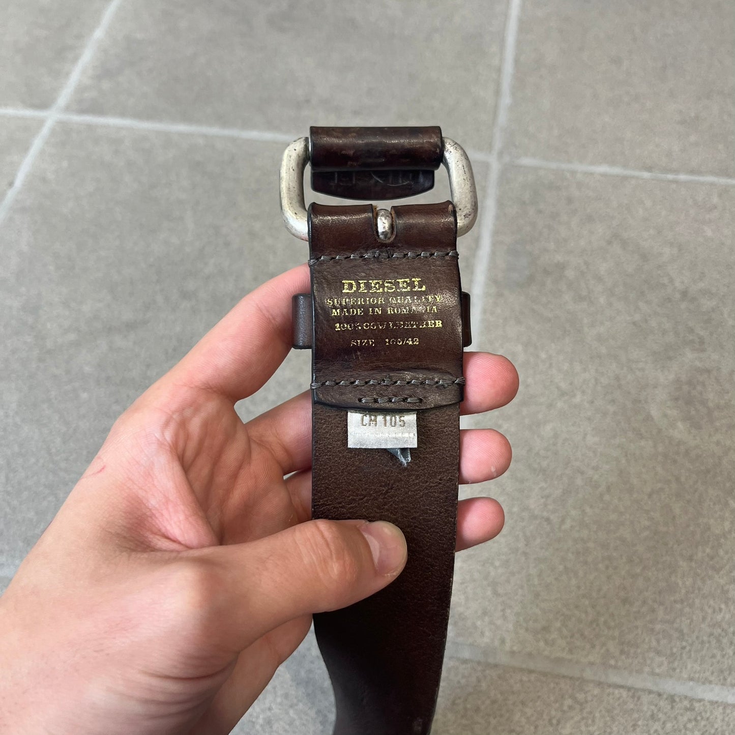Diesel 2000's Brown Leather Belt features a brown 100% cow leather construction with a simple silver-tone buckle. A small leather piece with Diesel text is attached to the buckle and rotates freely. The belt is marked “Extra Tough,” with gold Diesel text printed on the inside. Made in Italy, with an additional attached section at the end for adjusting the length.
