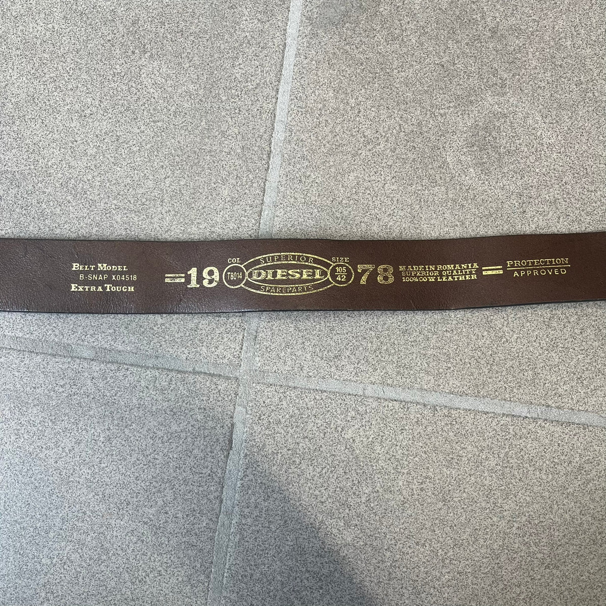Diesel 2000's Brown Leather Belt features a brown 100% cow leather construction with a simple silver-tone buckle. A small leather piece with Diesel text is attached to the buckle and rotates freely. The belt is marked “Extra Tough,” with gold Diesel text printed on the inside. Made in Italy, with an additional attached section at the end for adjusting the length.
