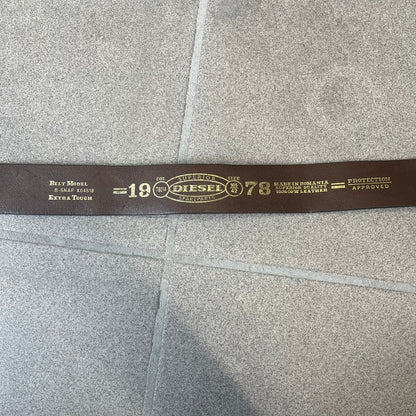 Diesel 2000's Brown Leather Belt features a brown 100% cow leather construction with a simple silver-tone buckle. A small leather piece with Diesel text is attached to the buckle and rotates freely. The belt is marked “Extra Tough,” with gold Diesel text printed on the inside. Made in Italy, with an additional attached section at the end for adjusting the length.
