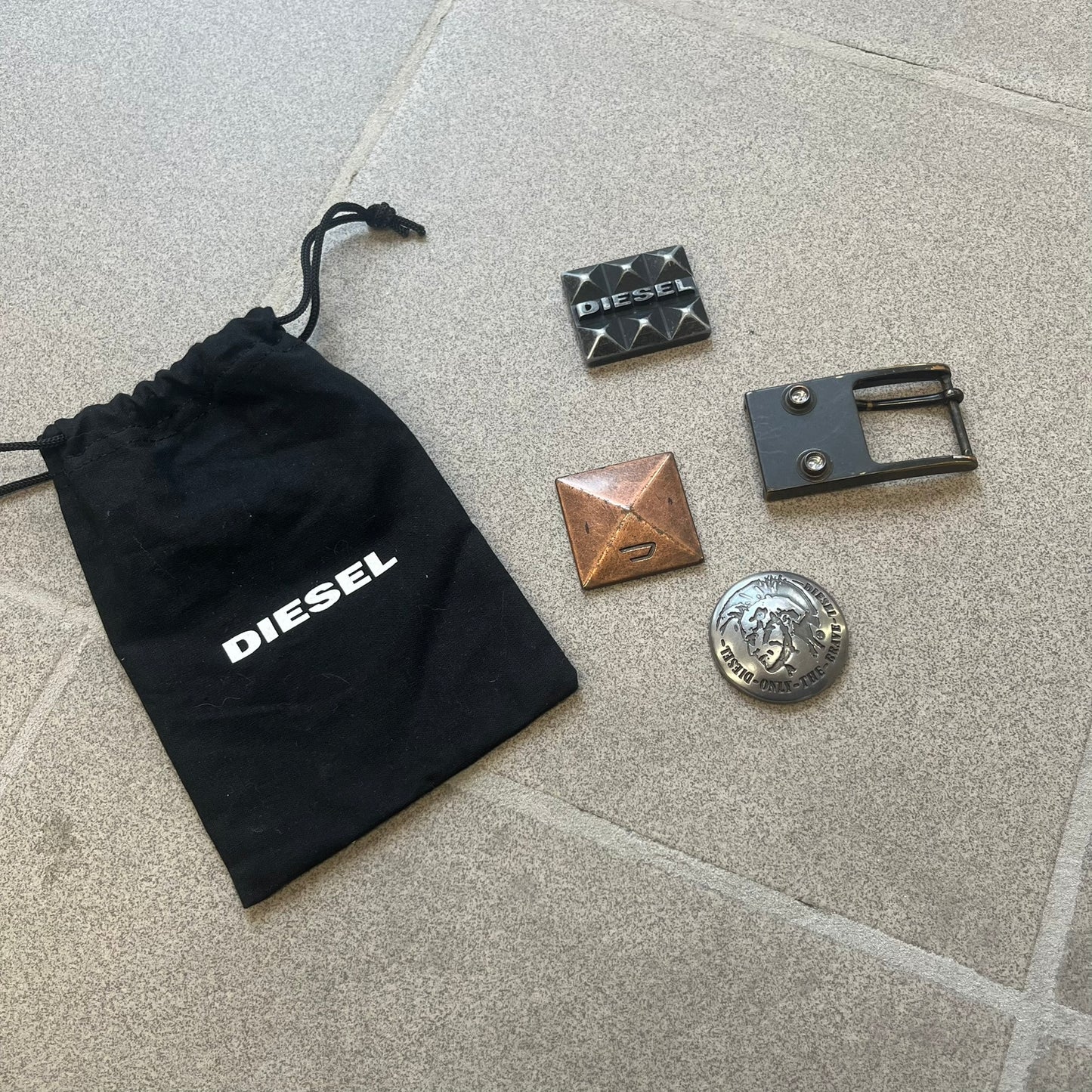 Diesel 2000s Changeable Buckles Set includes a Diesel belt buckle system with interchangeable front pieces. The base buckle features two snap buttons that allow different designs to be attached. The set includes three interchangeable buckles: a Diesel logo design with studs, a circular buckle with the Mohawk character logo, and a bronze pyramid logo design. Comes with the original dust bag.
