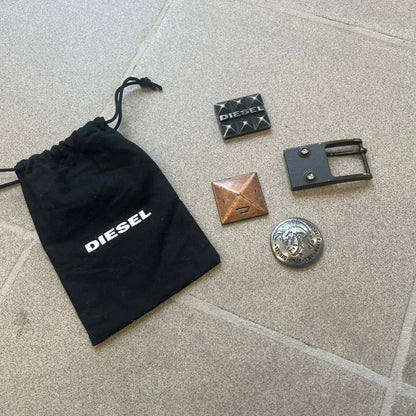 Diesel 2000s Changeable Buckles Set includes a Diesel belt buckle system with interchangeable front pieces. The base buckle features two snap buttons that allow different designs to be attached. The set includes three interchangeable buckles: a Diesel logo design with studs, a circular buckle with the Mohawk character logo, and a bronze pyramid logo design. Comes with the original dust bag.
