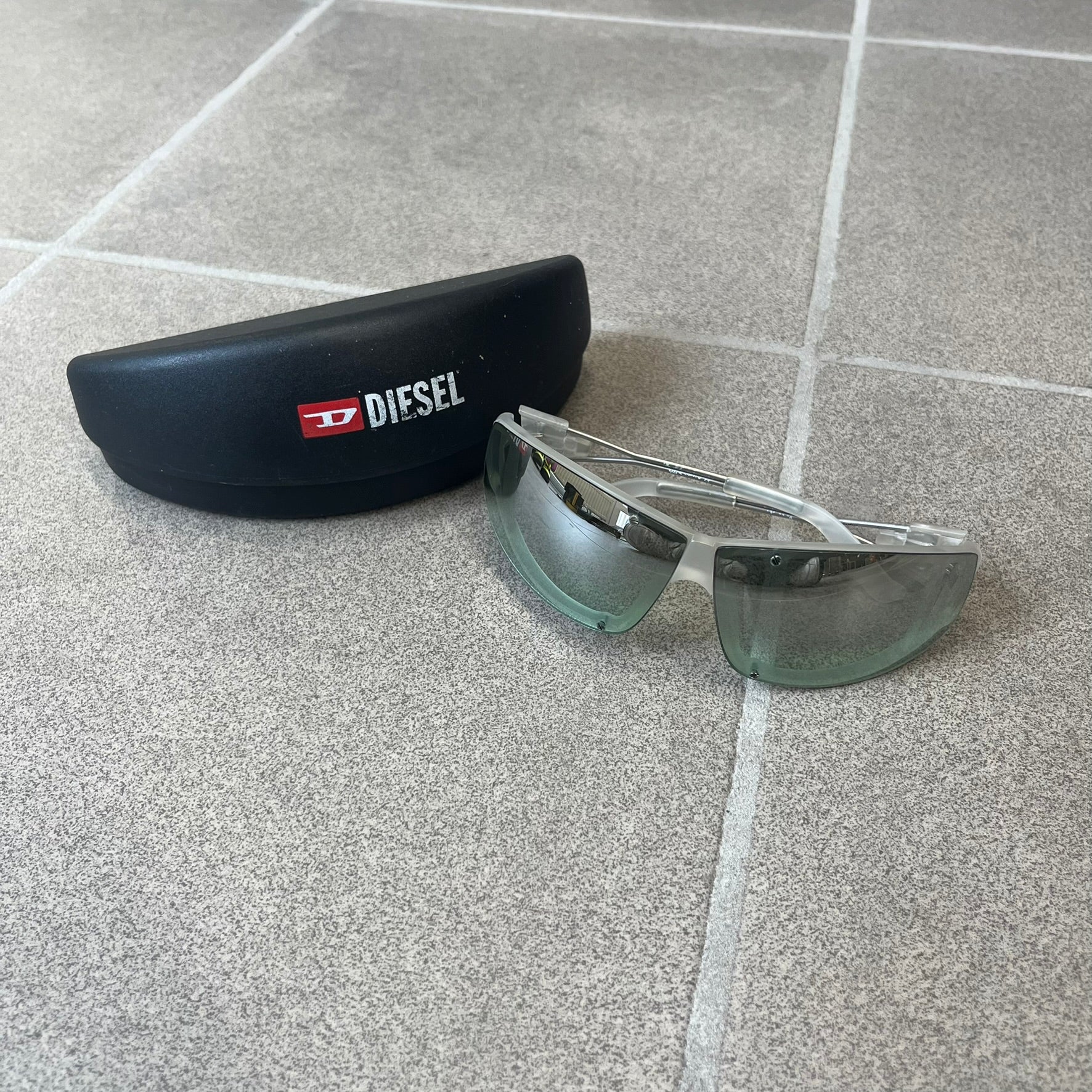 Diesel 2000's Clear Jumbo Sunglasses feature thin silver-tone wire temples with clear plastic sections extending over the ends and front of the frame. The lenses have a grey-tinted transparent finish, with visible screws on the front. A small silver Diesel logo is placed on the side, with white Diesel text printed in the corner of one lens.
