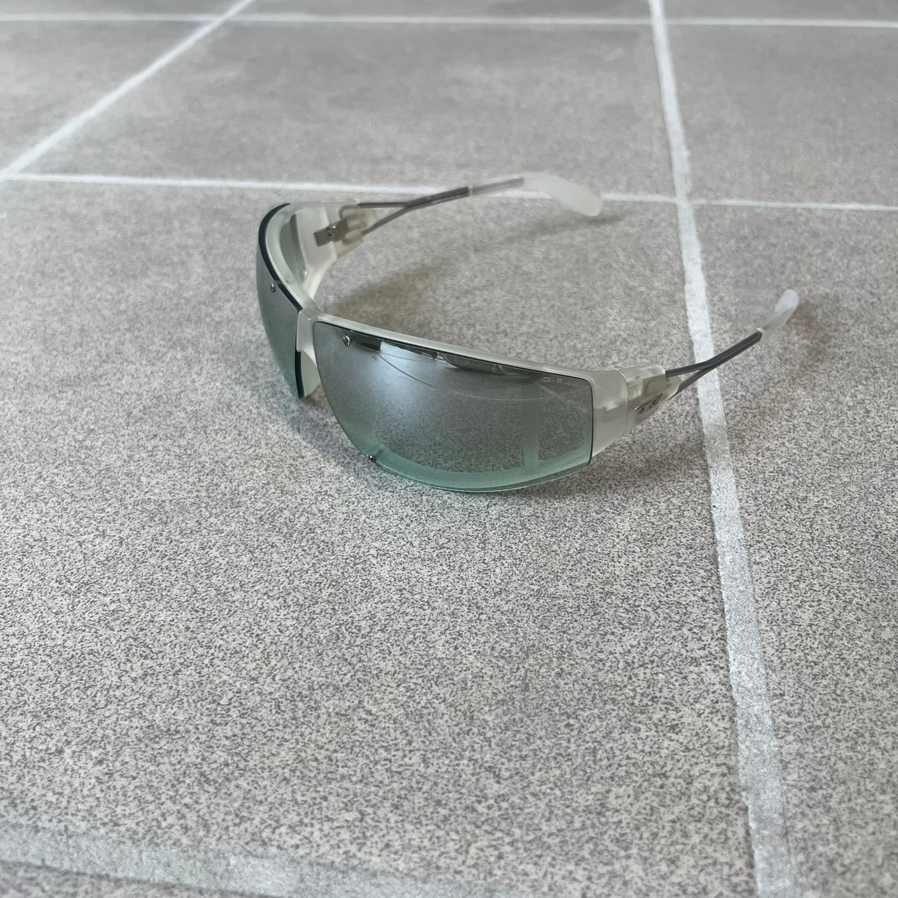 Diesel 2000's Clear Jumbo Sunglasses feature thin silver-tone wire temples with clear plastic sections extending over the ends and front of the frame. The lenses have a grey-tinted transparent finish, with visible screws on the front. A small silver Diesel logo is placed on the side, with white Diesel text printed in the corner of one lens.
