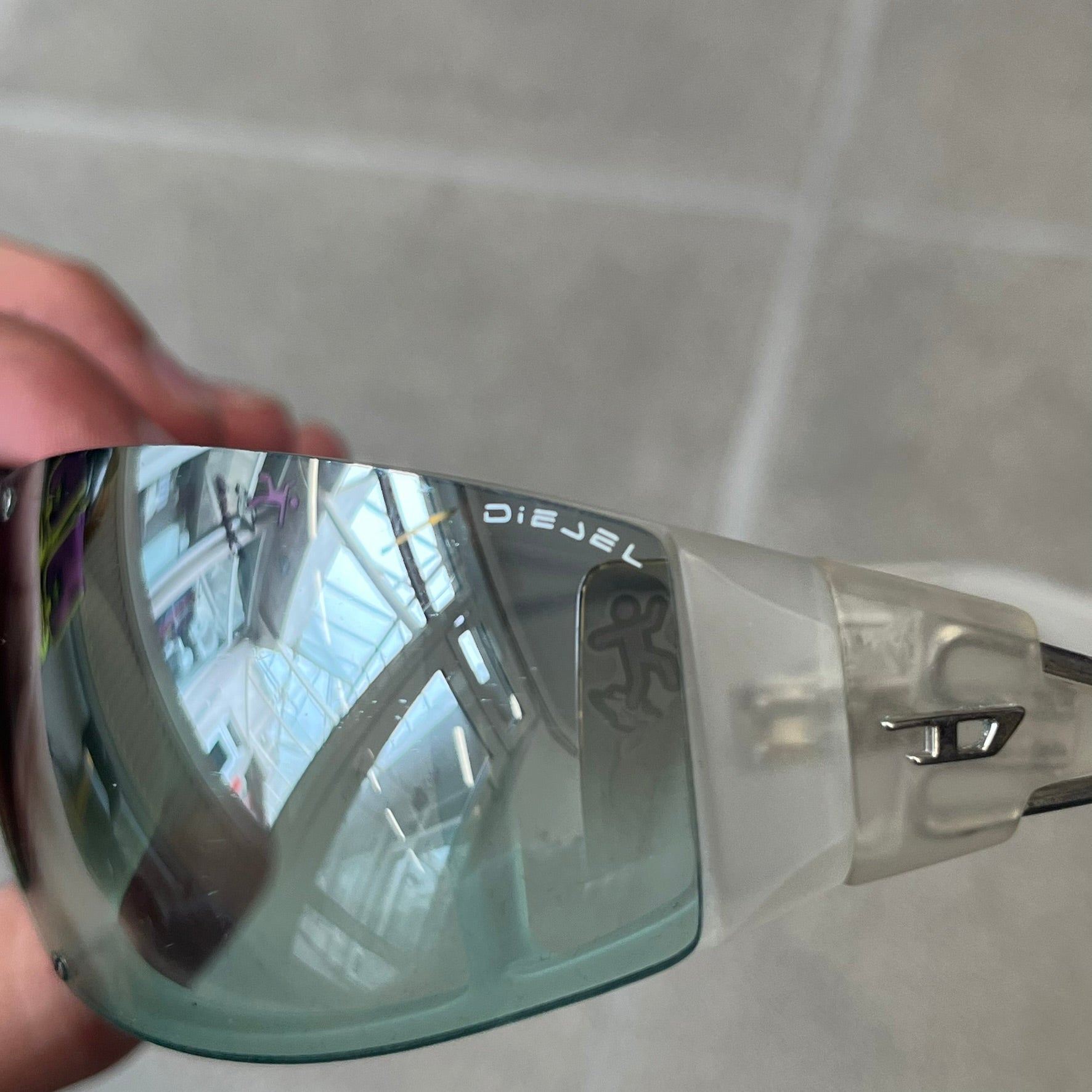 Diesel 2000's Clear Jumbo Sunglasses feature thin silver-tone wire temples with clear plastic sections extending over the ends and front of the frame. The lenses have a grey-tinted transparent finish, with visible screws on the front. A small silver Diesel logo is placed on the side, with white Diesel text printed in the corner of one lens.
