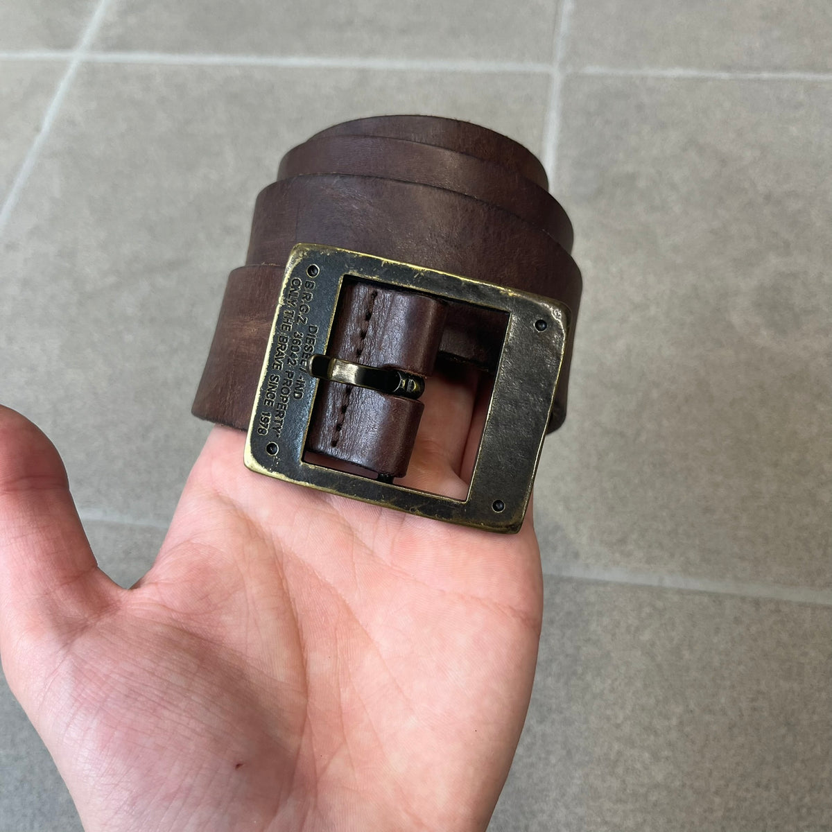 Diesel 2000's Only The Brave Brown Leather Belt features a brown 100% cow leather construction with a bronze-tone buckle. The buckle has engraved Diesel branding, including “Only The Brave Since 1978” text and other details. The belt is marked “Extra Tough,” with gold Diesel text printed on the inside. Made in Italy.
