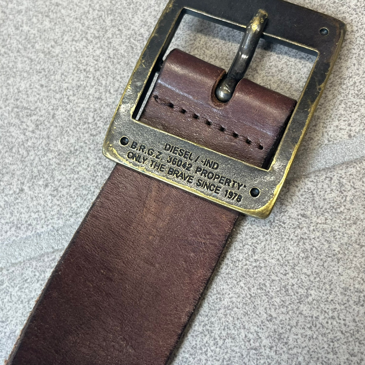 Diesel 2000's Only The Brave Brown Leather Belt features a brown 100% cow leather construction with a bronze-tone buckle. The buckle has engraved Diesel branding, including “Only The Brave Since 1978” text and other details. The belt is marked “Extra Tough,” with gold Diesel text printed on the inside. Made in Italy.
