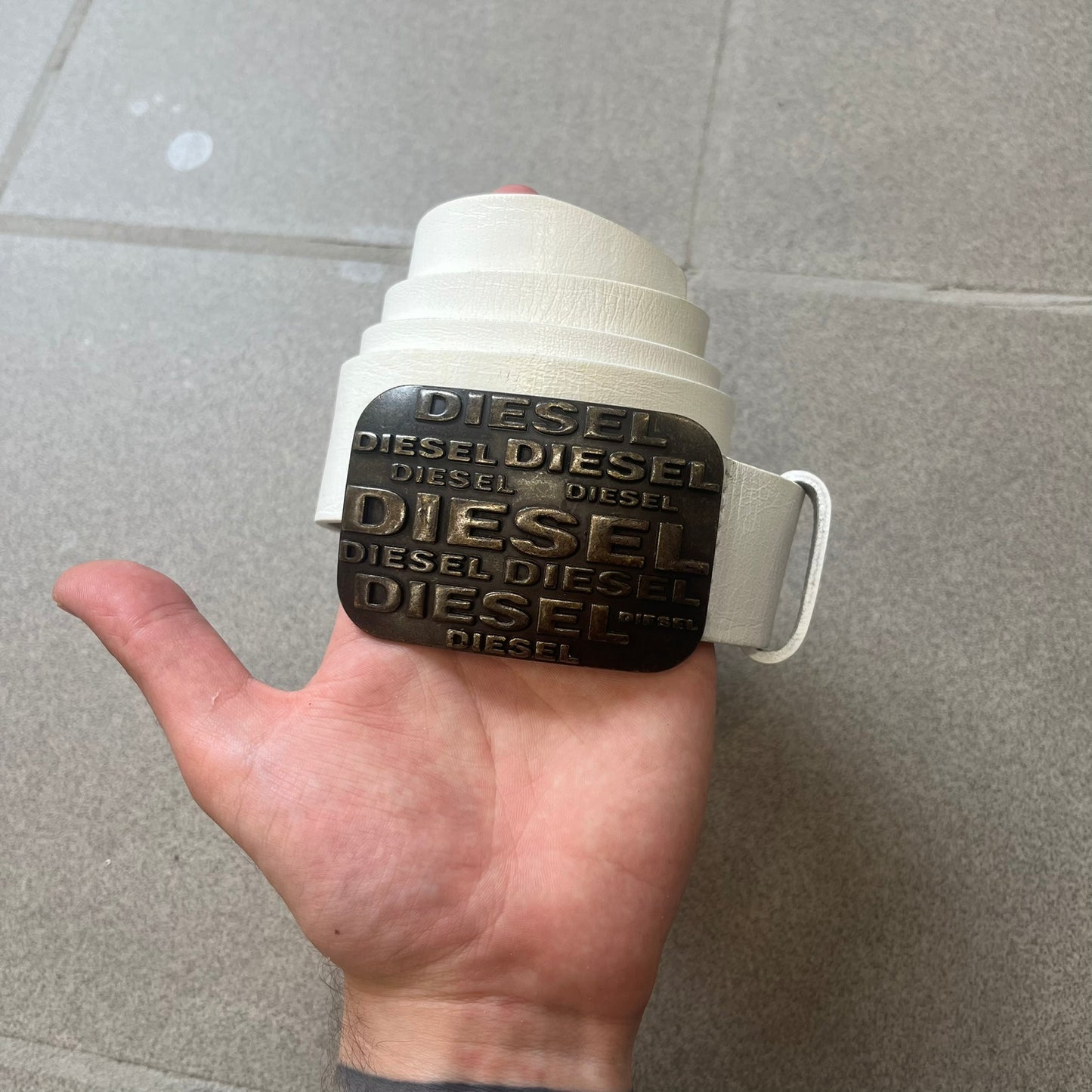 Diesel 2000s Repeat Logo White Leather Belt features a white leather strap with a rectangular bronze-tone buckle. The buckle is decorated with a repeating Diesel text logo pattern in different sizes, creating a layered branded design. Made in Italy.
