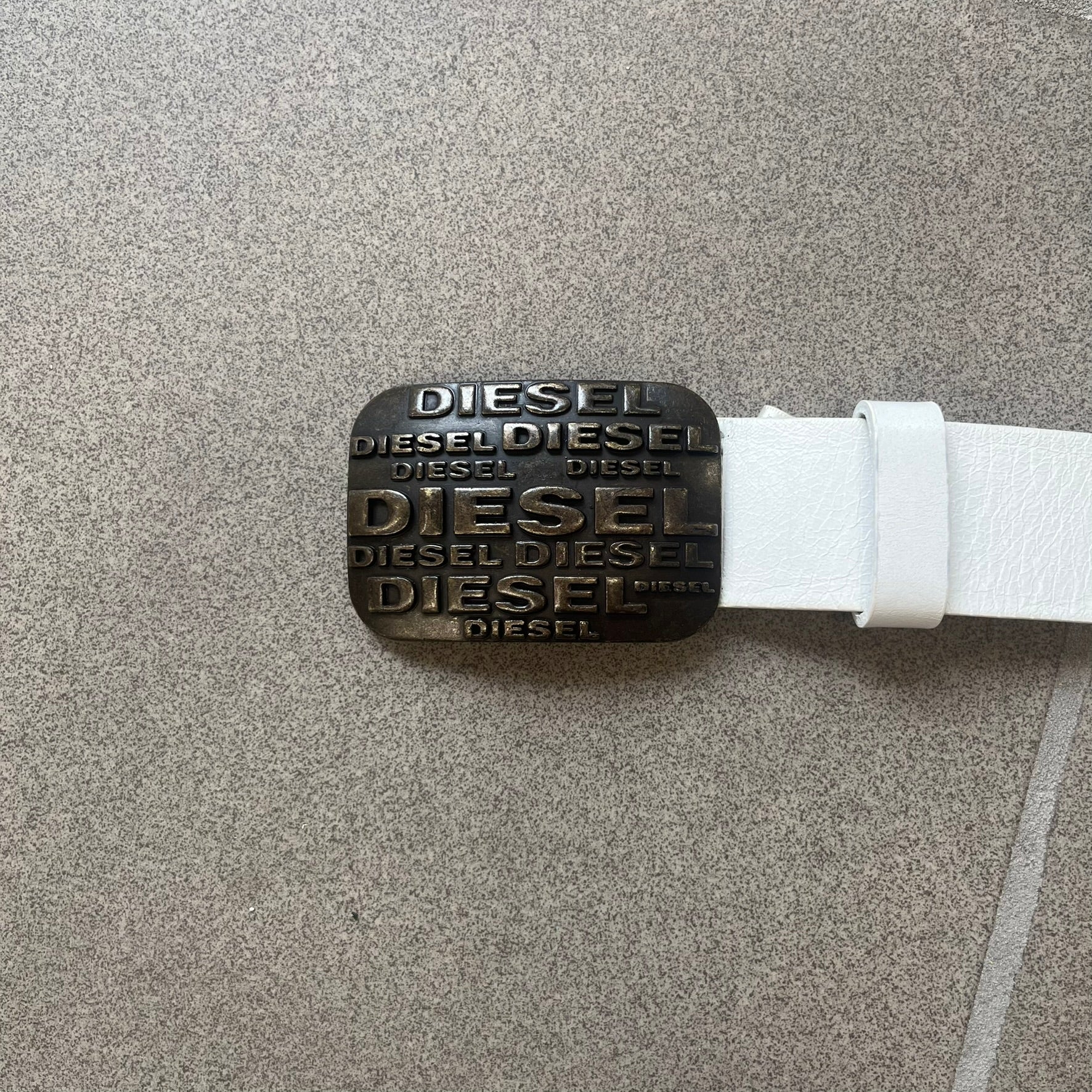Diesel 2000s Repeat Logo White Leather Belt features a white leather strap with a rectangular bronze-tone buckle. The buckle is decorated with a repeating Diesel text logo pattern in different sizes, creating a layered branded design. Made in Italy.
