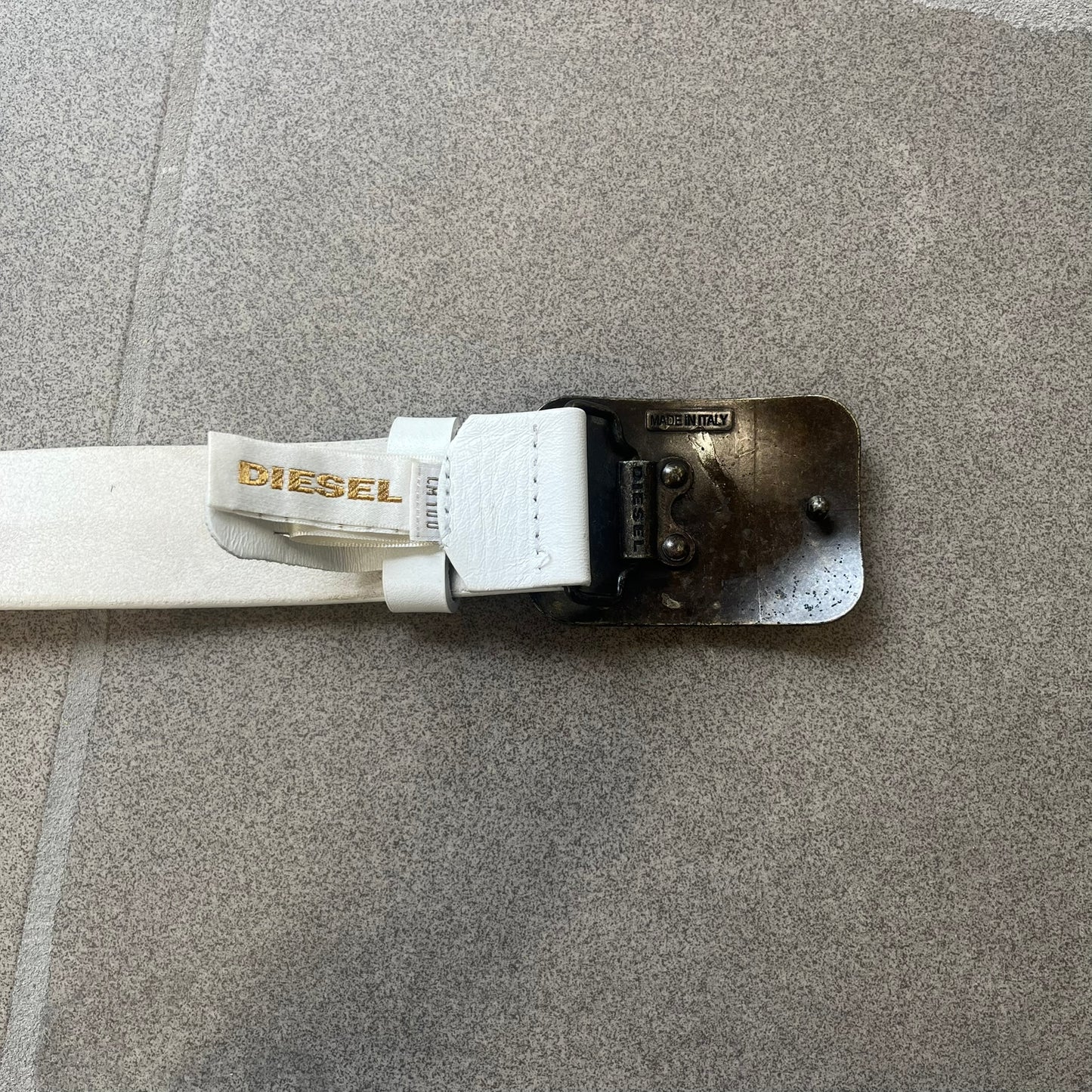 Diesel 2000s Repeat Logo White Leather Belt features a white leather strap with a rectangular bronze-tone buckle. The buckle is decorated with a repeating Diesel text logo pattern in different sizes, creating a layered branded design. Made in Italy.
