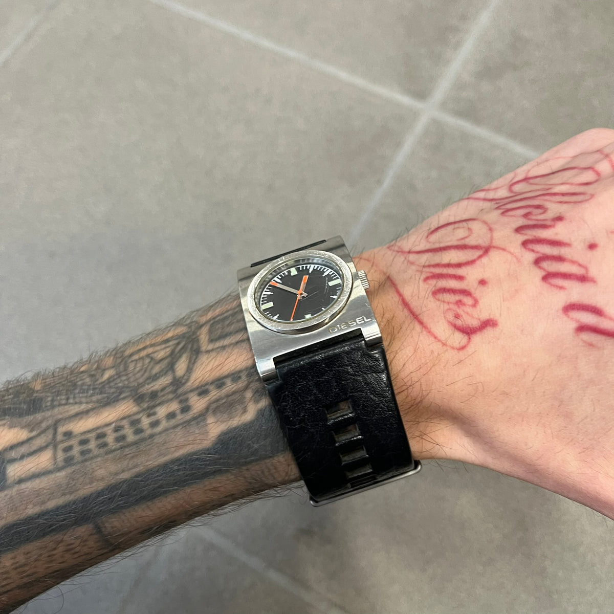 Diesel 2000’s Square DZ2107 Analog Watch has a square stainless steel case with an engraved Diesel logo in the bottom right corner. The circular black dial has orange hands, and the watch is fitted with a black leather strap.