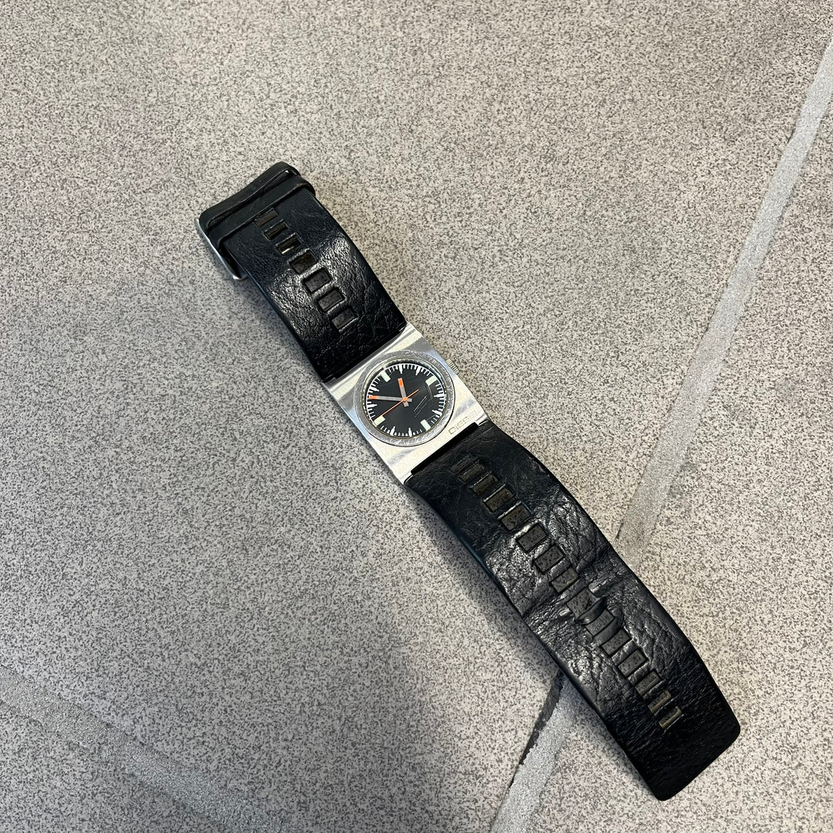 Diesel 2000’s Square DZ2107 Analog Watch has a square stainless steel case with an engraved Diesel logo in the bottom right corner. The circular black dial has orange hands, and the watch is fitted with a black leather strap.