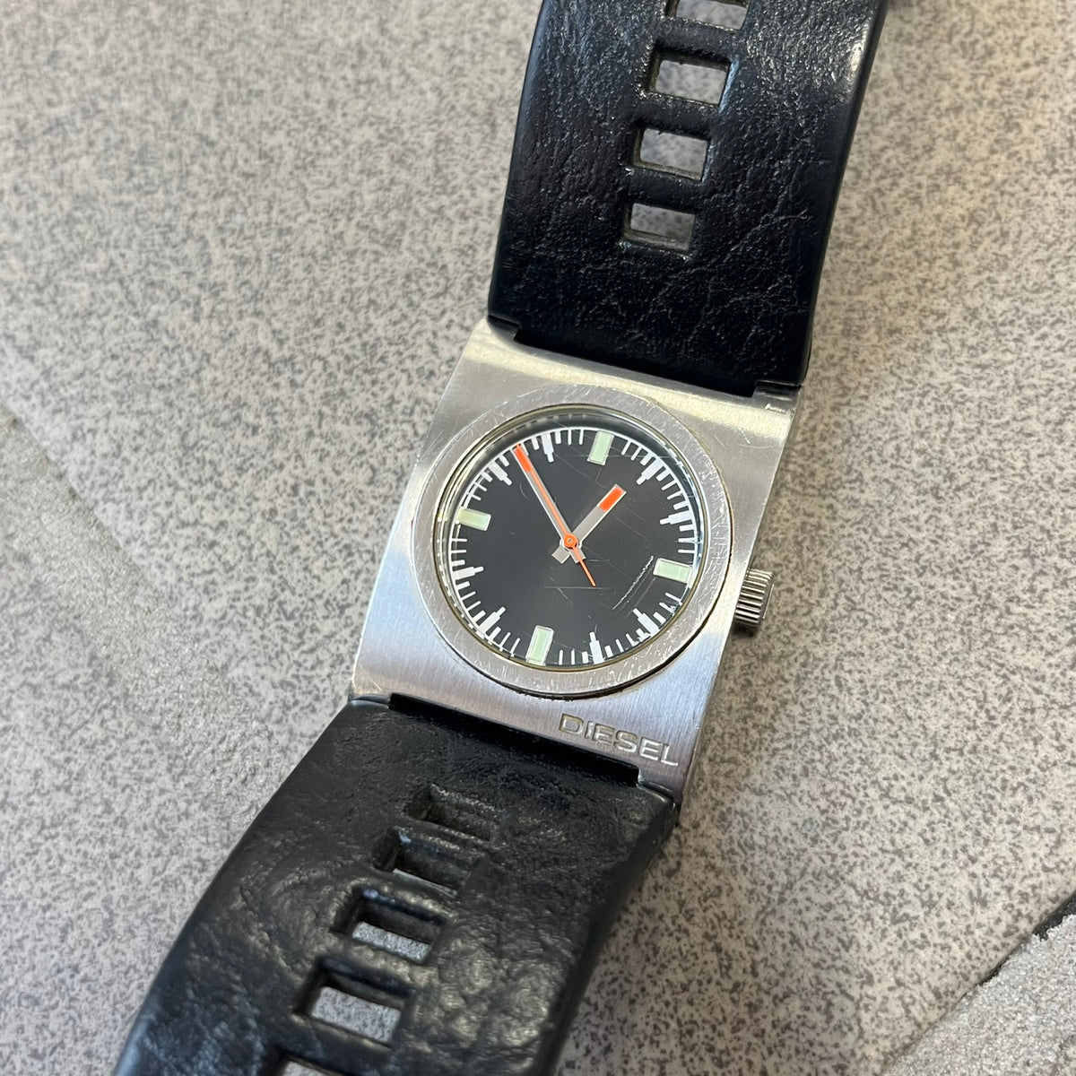 Diesel 2000’s Square DZ2107 Analog Watch has a square stainless steel case with an engraved Diesel logo in the bottom right corner. The circular black dial has orange hands, and the watch is fitted with a black leather strap.