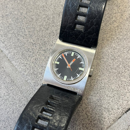 Diesel 2000’s Square DZ2107 Analog Watch has a square stainless steel case with an engraved Diesel logo in the bottom right corner. The circular black dial has orange hands, and the watch is fitted with a black leather strap.