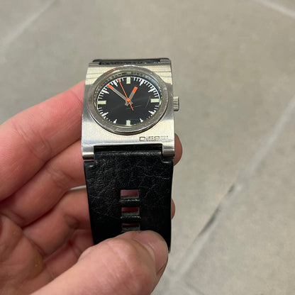 Diesel 2000’s Square DZ2107 Analog Watch has a square stainless steel case with an engraved Diesel logo in the bottom right corner. The circular black dial has orange hands, and the watch is fitted with a black leather strap.