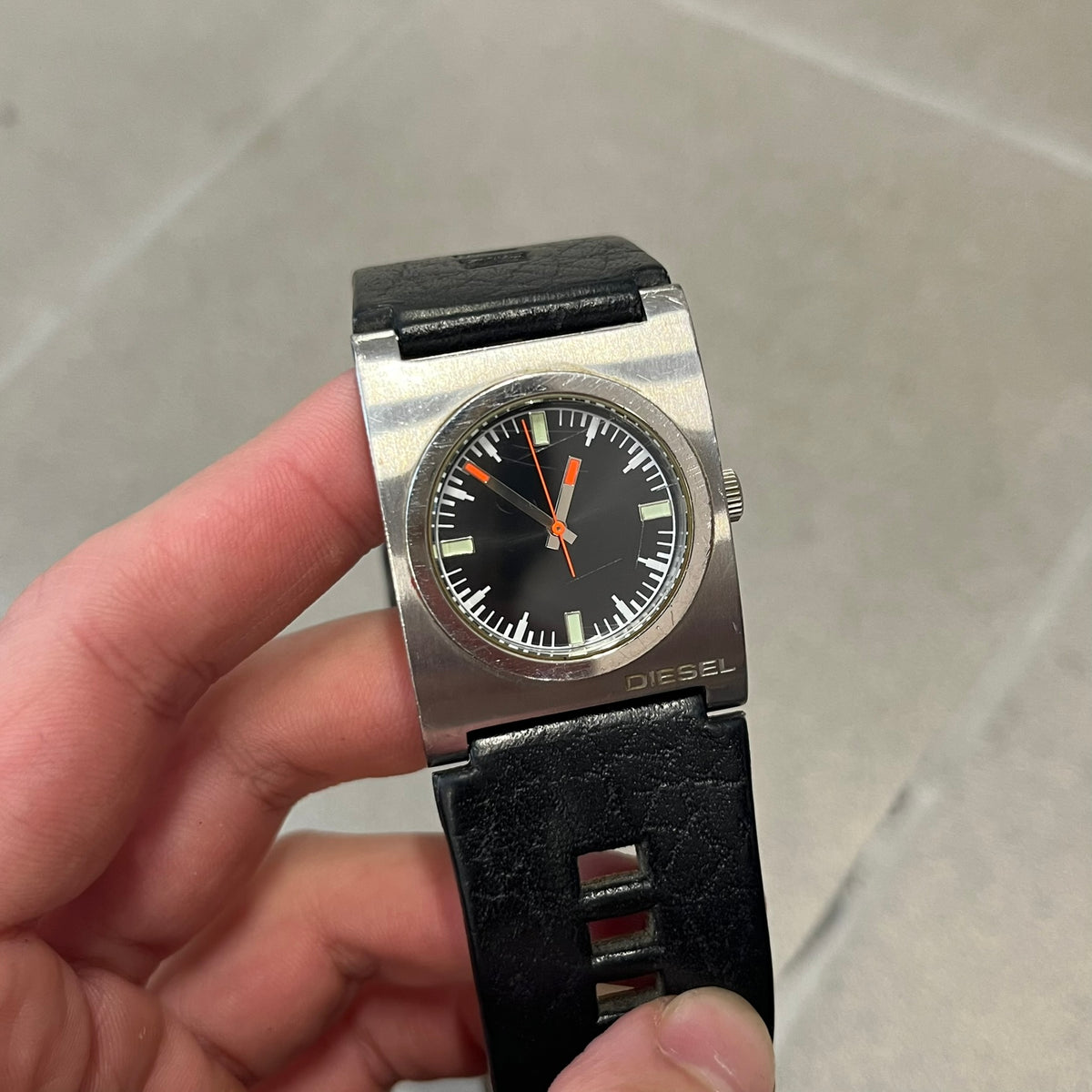 Diesel 2000’s Square DZ2107 Analog Watch has a square stainless steel case with an engraved Diesel logo in the bottom right corner. The circular black dial has orange hands, and the watch is fitted with a black leather strap.