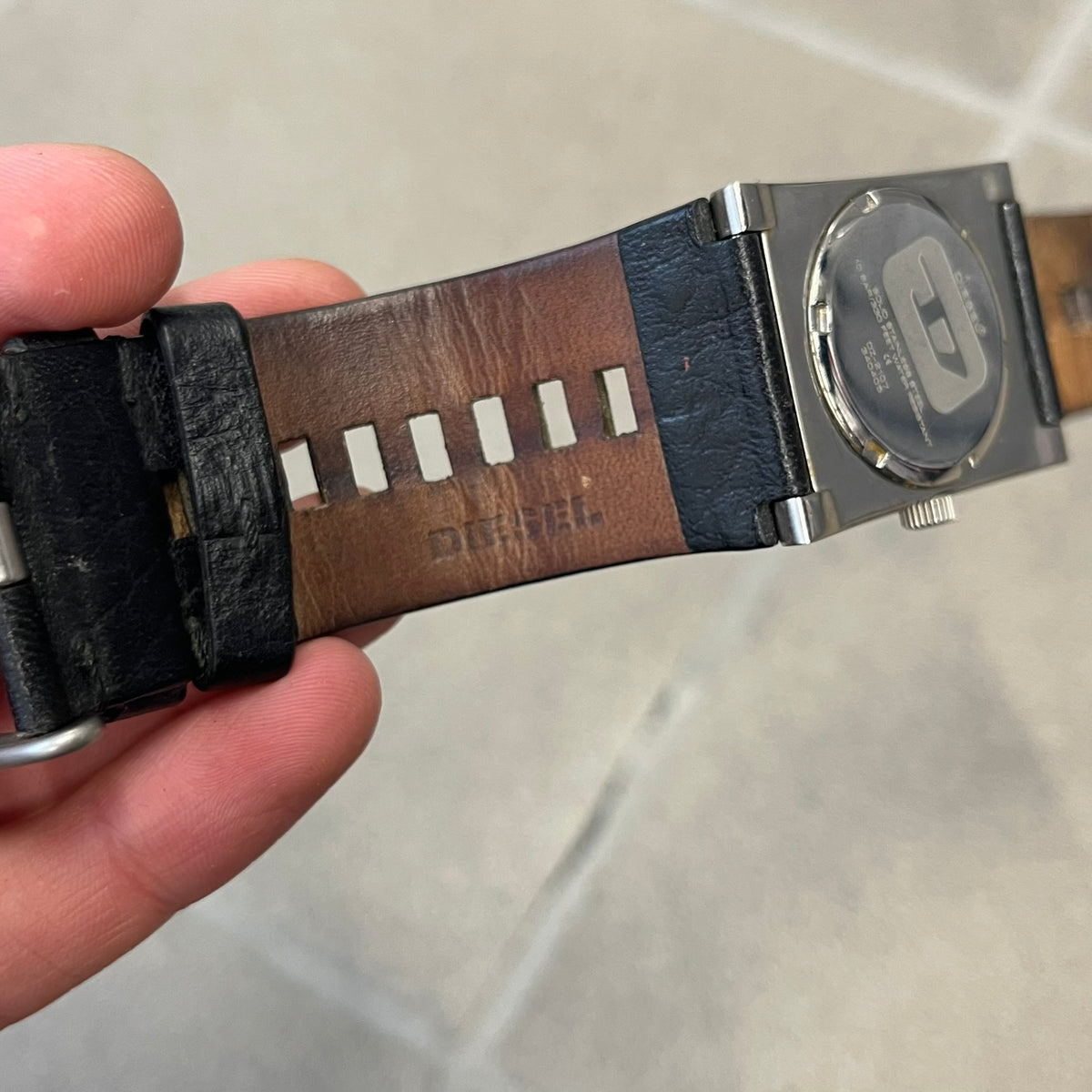 Diesel 2000’s Square DZ2107 Analog Watch has a square stainless steel case with an engraved Diesel logo in the bottom right corner. The circular black dial has orange hands, and the watch is fitted with a black leather strap.
