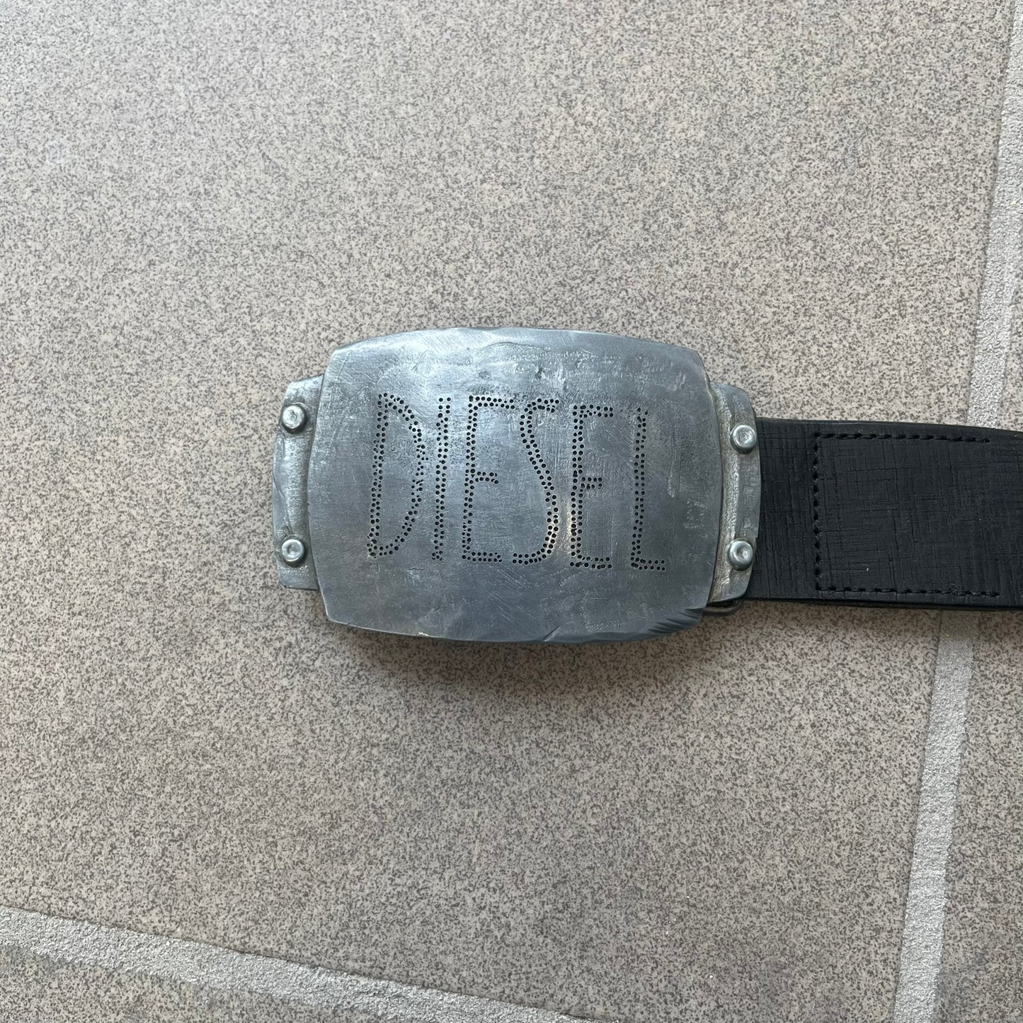 Diesel 2000s Vintage Buckle Leather Belt features a black leather strap made in Italy. The belt has a large rectangular metal buckle designed to look like a worn metal sheet, with a Diesel logo formed from small perforated holes. The buckle includes visible screws on the sides and the belt is made from 100% leather.

