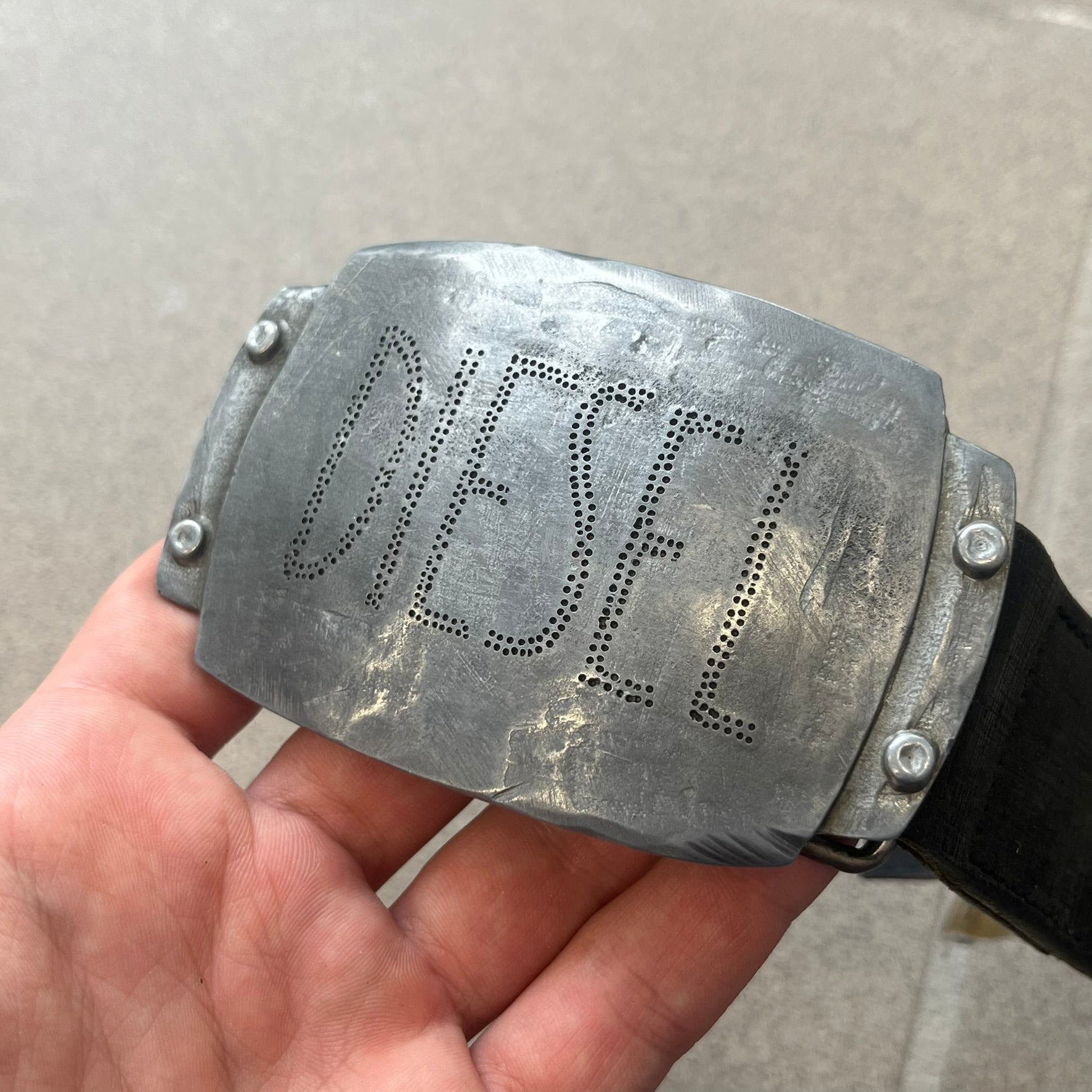 Diesel 2000s Vintage Buckle Leather Belt features a black leather strap made in Italy. The belt has a large rectangular metal buckle designed to look like a worn metal sheet, with a Diesel logo formed from small perforated holes. The buckle includes visible screws on the sides and the belt is made from 100% leather.
