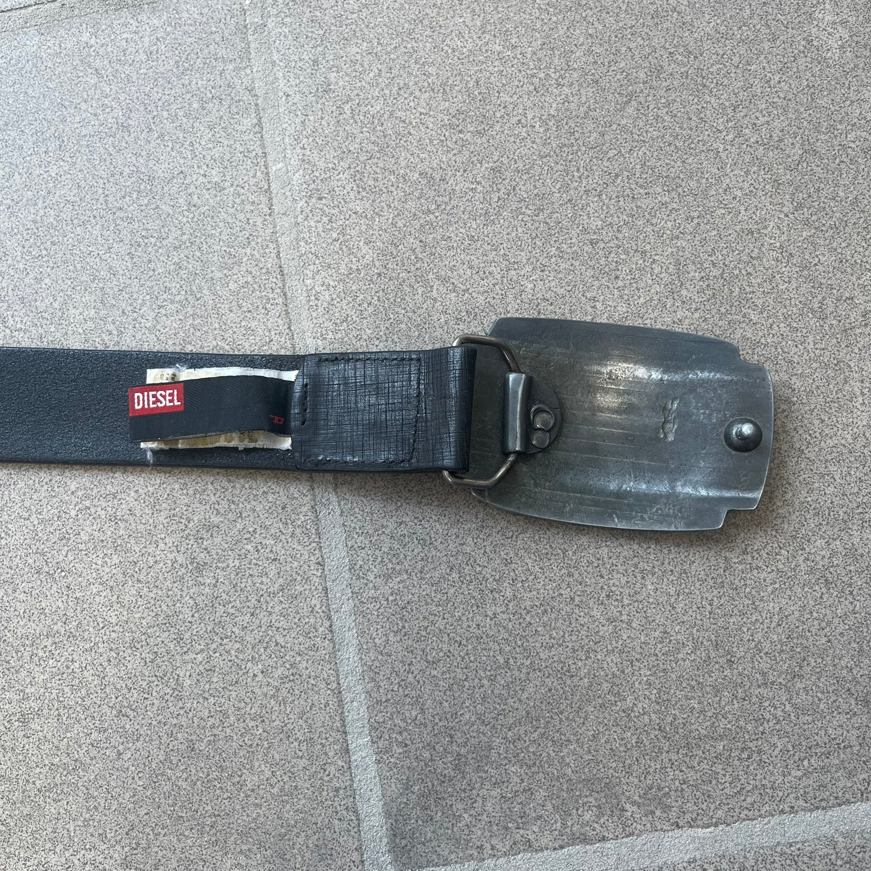 Diesel 2000s Vintage Buckle Leather Belt features a black leather strap made in Italy. The belt has a large rectangular metal buckle designed to look like a worn metal sheet, with a Diesel logo formed from small perforated holes. The buckle includes visible screws on the sides and the belt is made from 100% leather.
